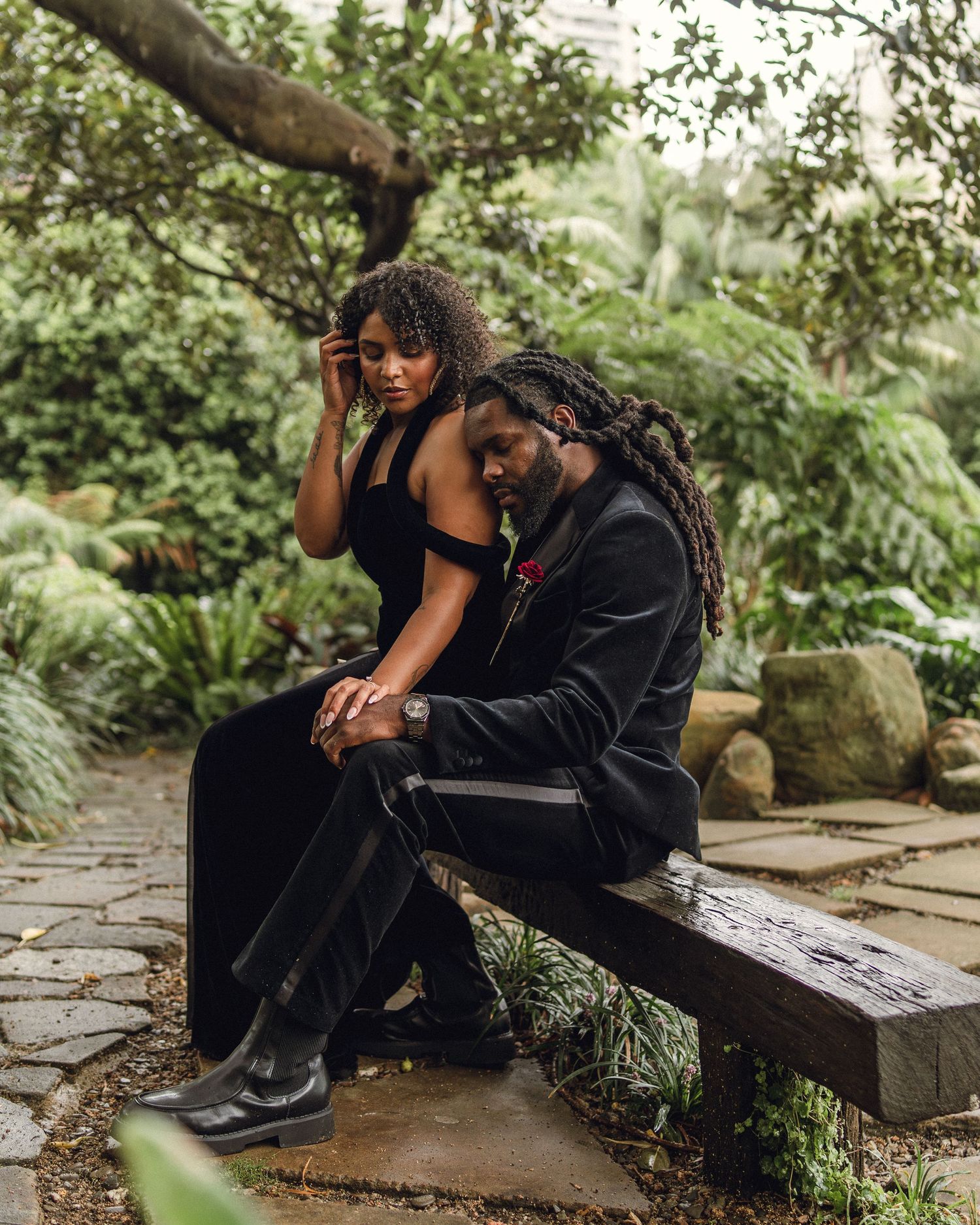 Stylish couple in black outfits sitting together on a bench in a garden.