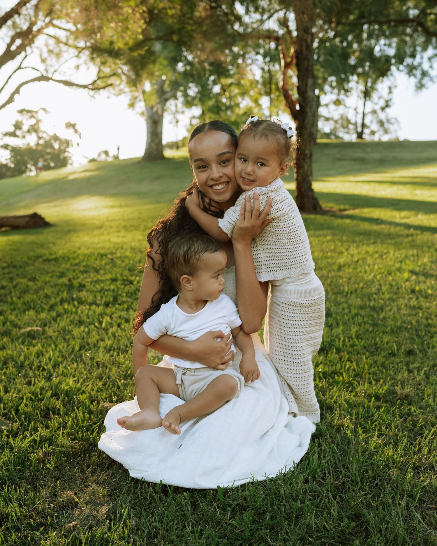 A smiling mother sits on grass with her two young children hugging her in a sunny park.