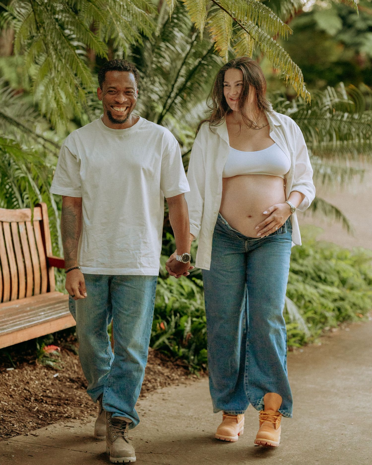 Smiling couple walking hand in hand on a path, pregnant woman cradling her baby bump in a tropical garden.