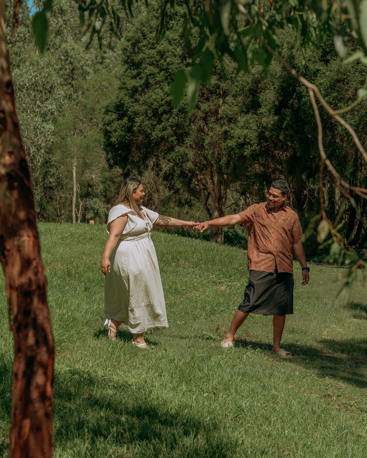 Couple holding hands and dancing together on green grass surrounded by eucalyptus trees in a sunny park.