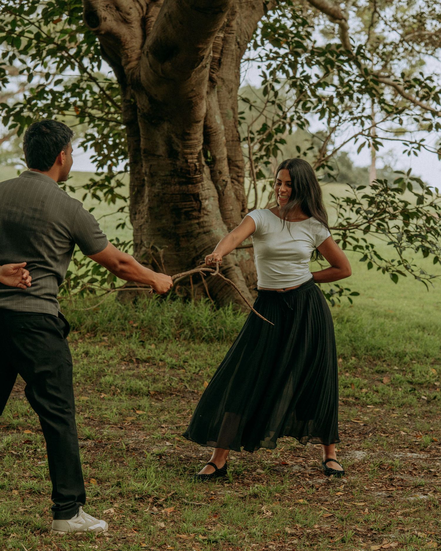 A smiling woman in a black skirt holds a stick while playfully interacting with her partner near a large tree.