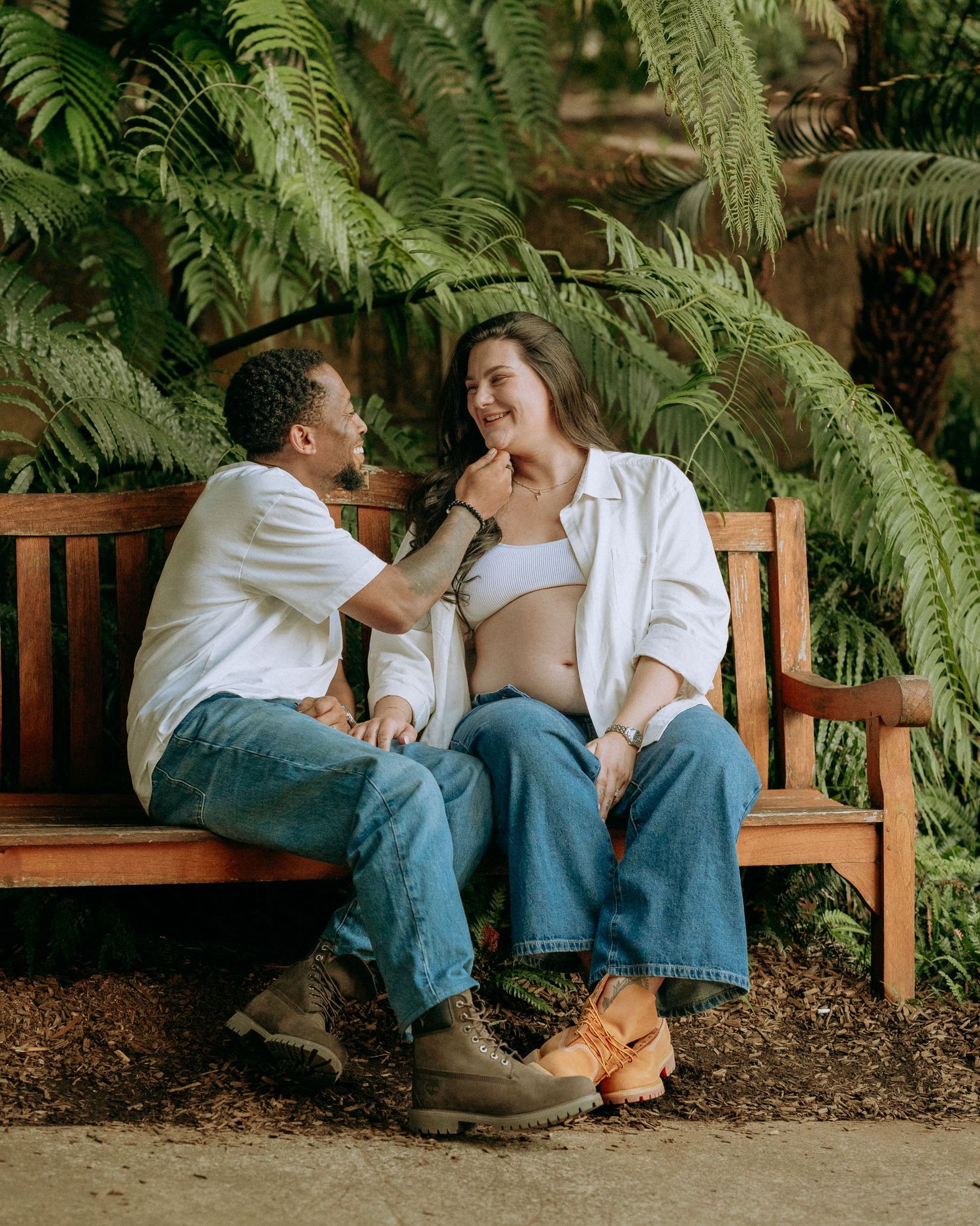 Expecting couple sitting on a wooden bench surrounded by ferns, sharing a tender moment together.