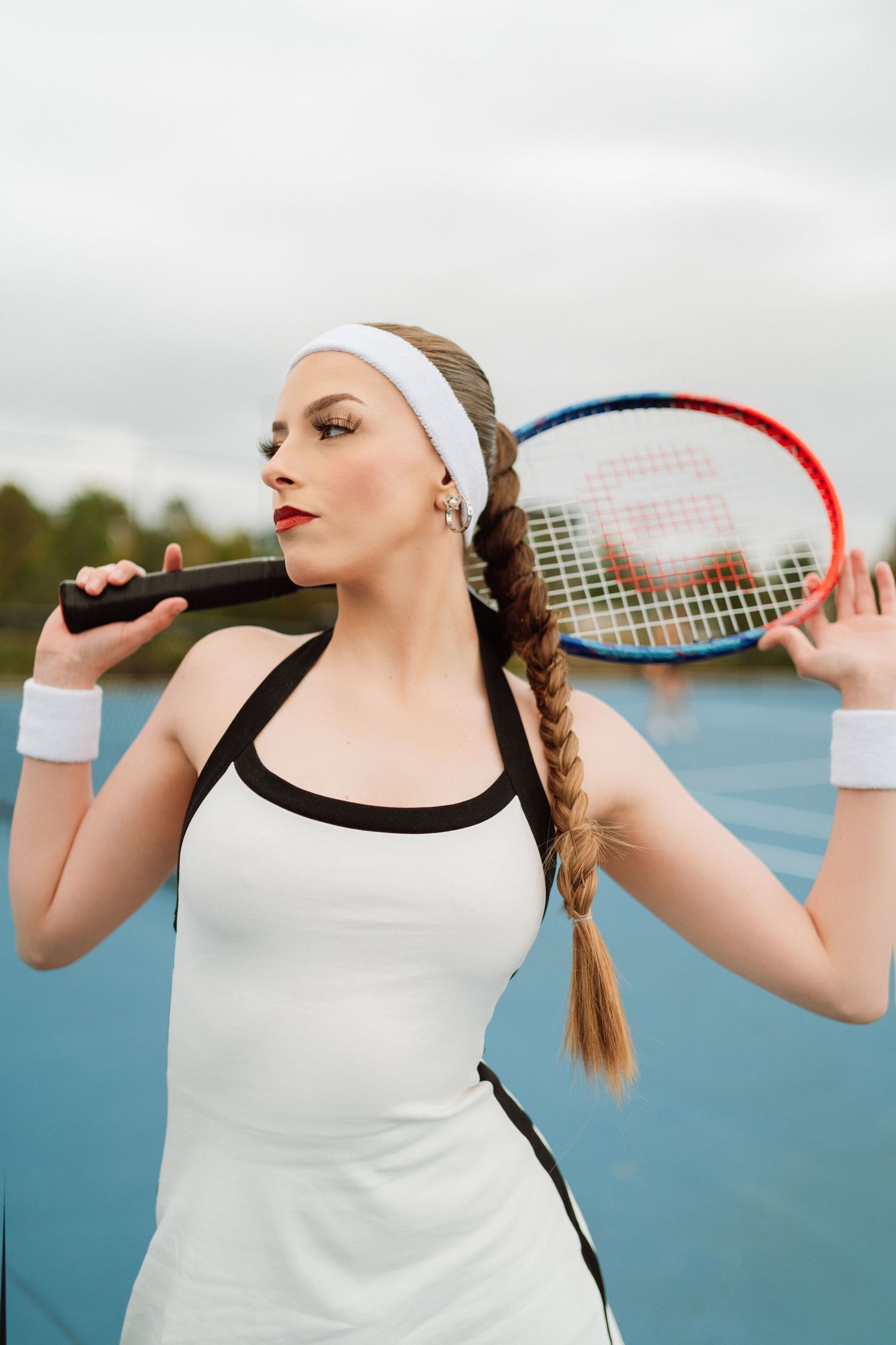 Young woman in white tennis dress holding Wilson racket on blue outdoor court.