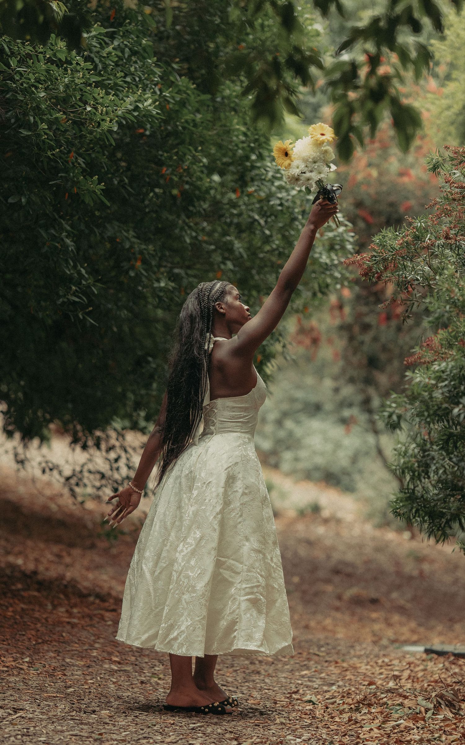 A young Black woman in a white dress reaches up to hold a yellow bouquet among lush green trees outdoors.