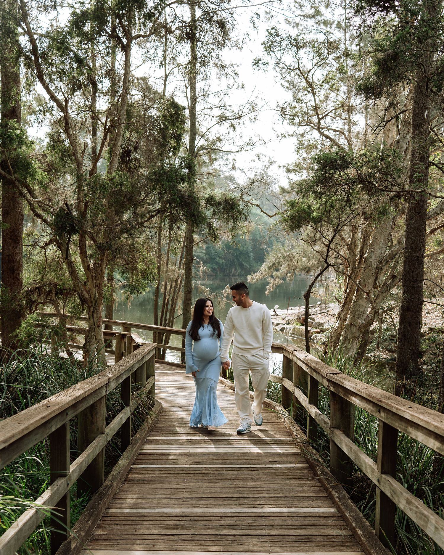 Couple walking on a wooden boardwalk through lush forest, woman in blue maternity dress, man in white outfit.