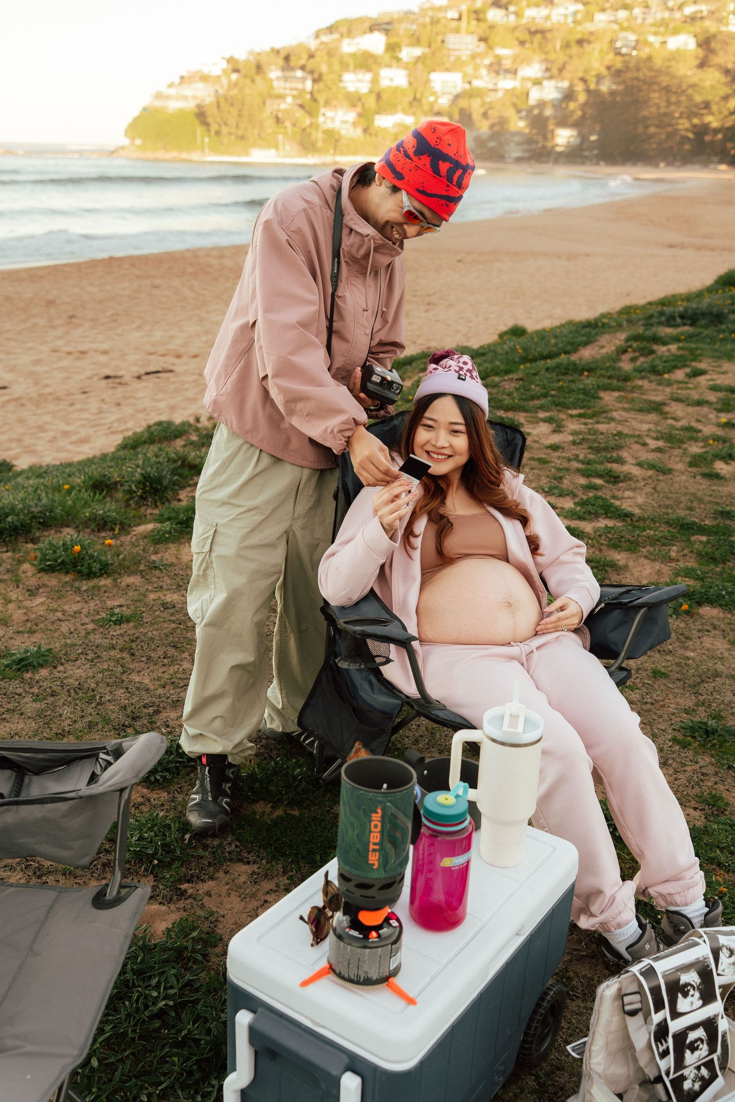 Pregnant woman relaxes in beach chair while partner tends to her at a cozy seaside campsite.