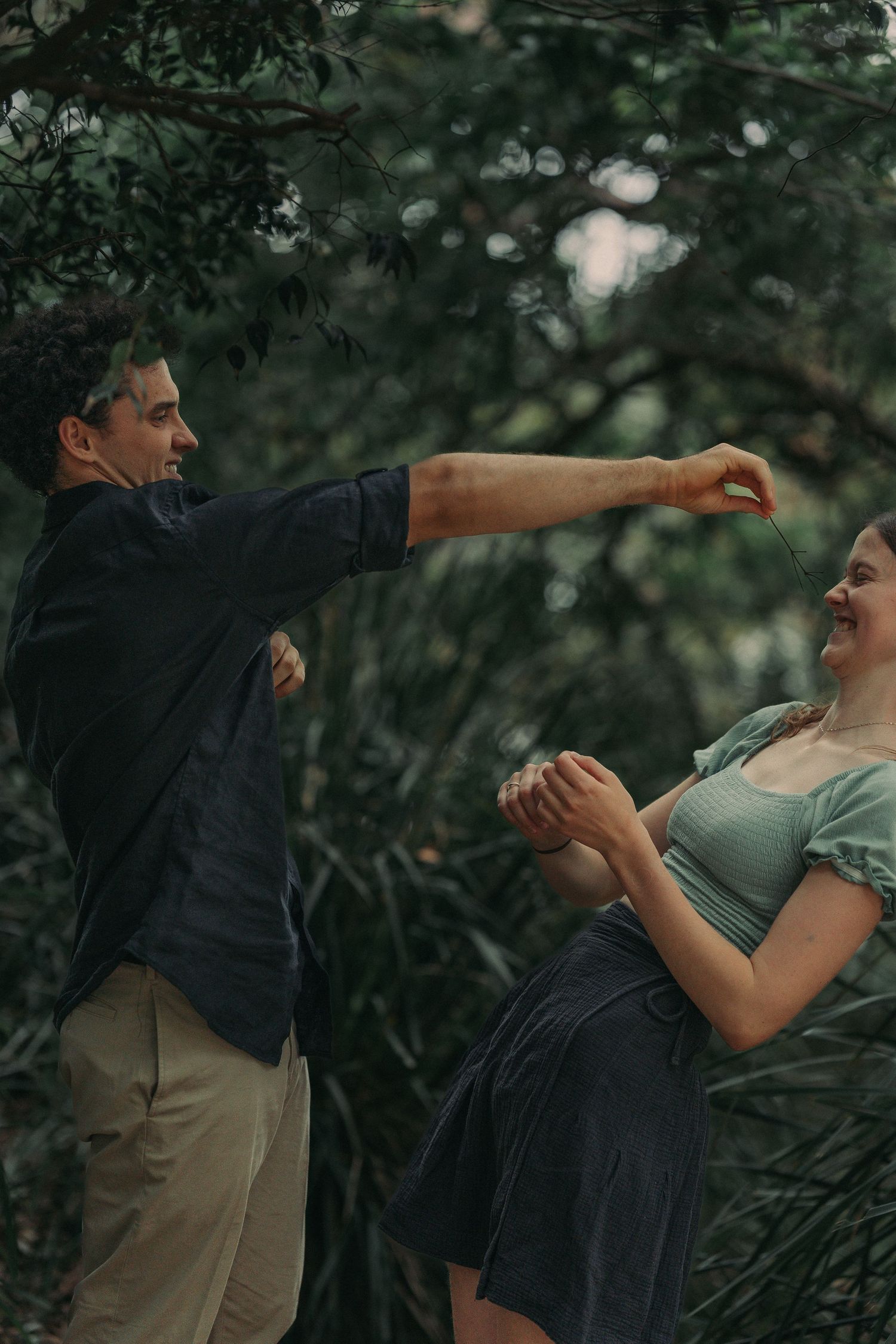 A joyful couple dancing and laughing together surrounded by lush green trees.