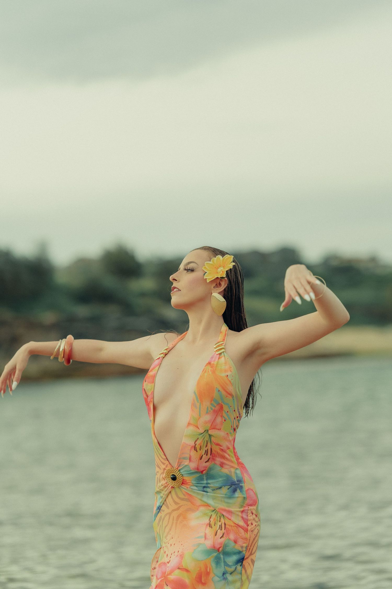 Woman in colorful tropical halter dress with yellow flower in hair posing gracefully by water.