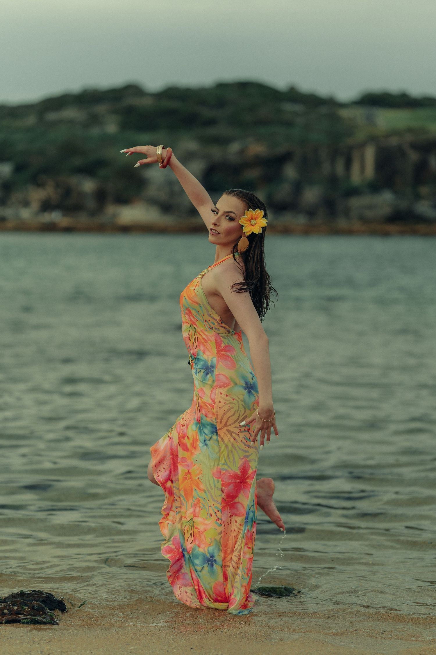 Woman in vibrant tropical floral dress dancing gracefully in ocean water with one arm raised at beach.