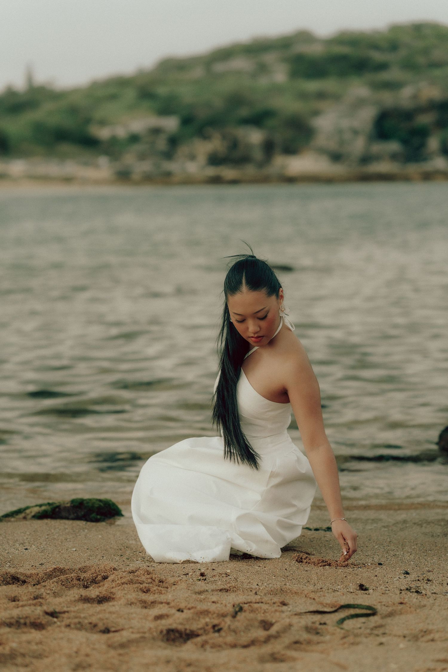 Young woman in white dress kneeling on sandy beach touching the sand with water and hills behind.