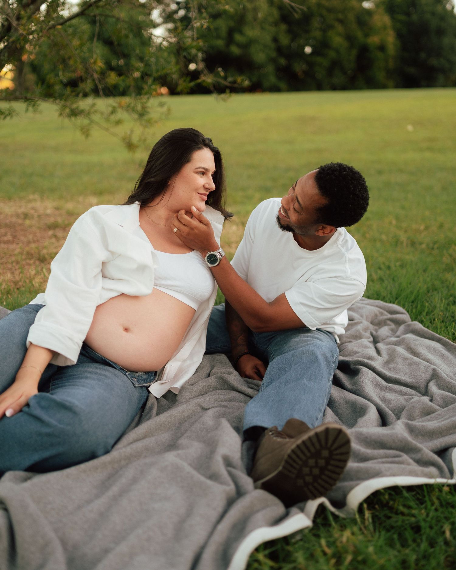Interracial couple sitting on a blanket outdoors, smiling at each other during a maternity shoot.