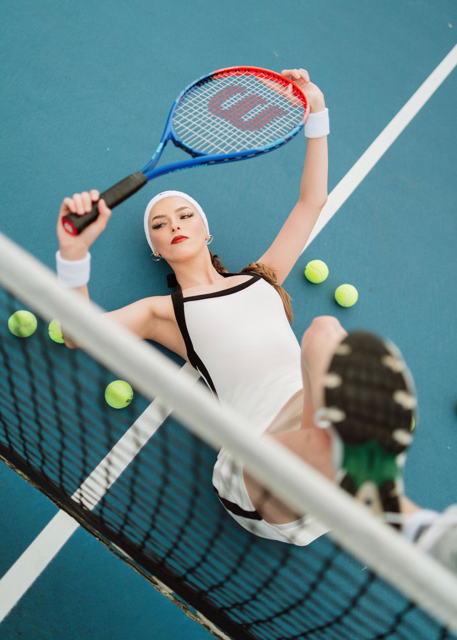 Female tennis player in white outfit poses dramatically on blue court with Wilson racket and tennis balls.