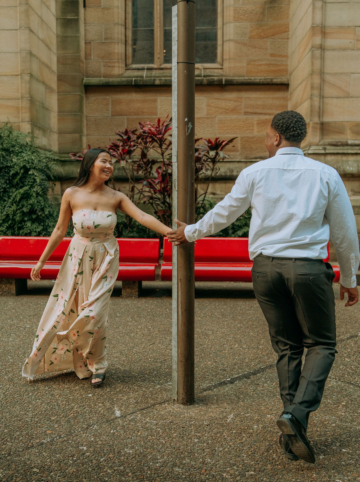 Couple playfully holding hands around a pole in a courtyard near a sandstone building.