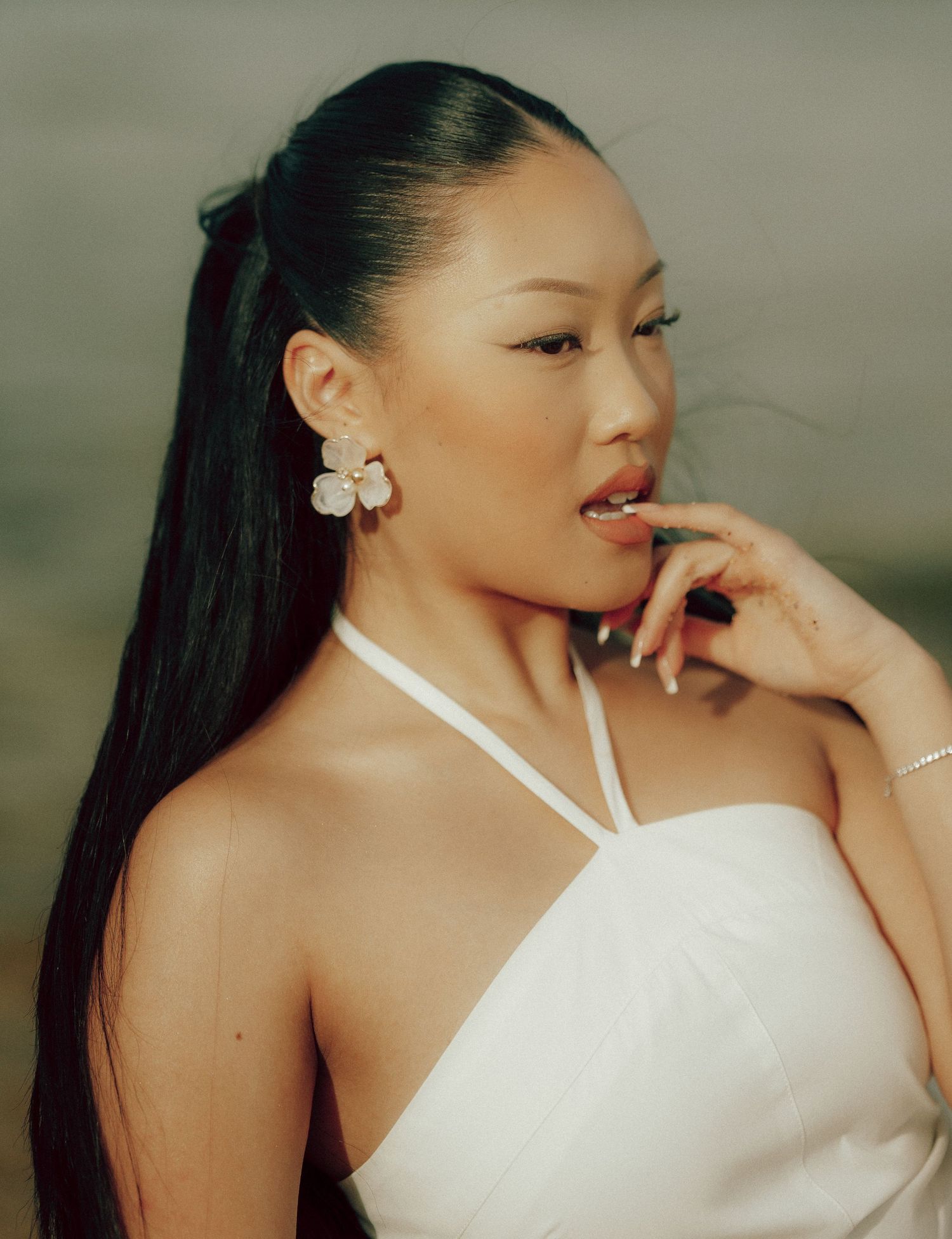 Side profile portrait of Asian woman in white top wearing large floral earrings with sleek ponytail outdoors.