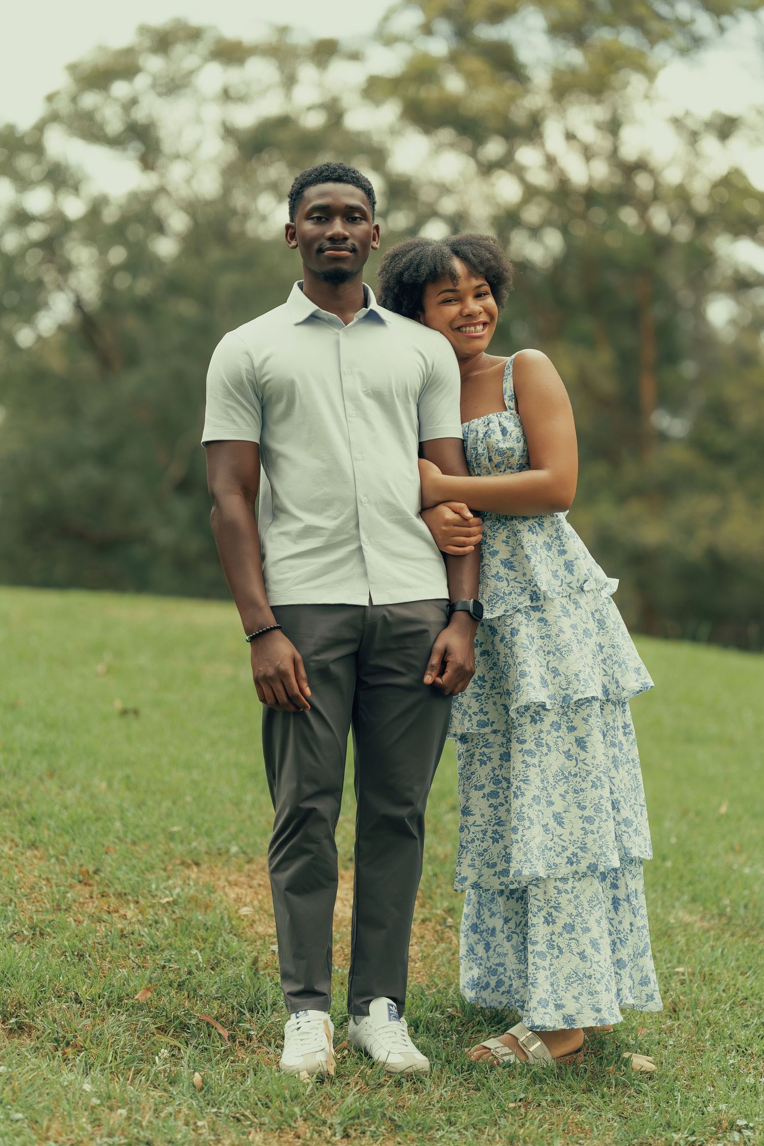 A couple poses together on green grass in a park, the woman in a floral dress smiling warmly.