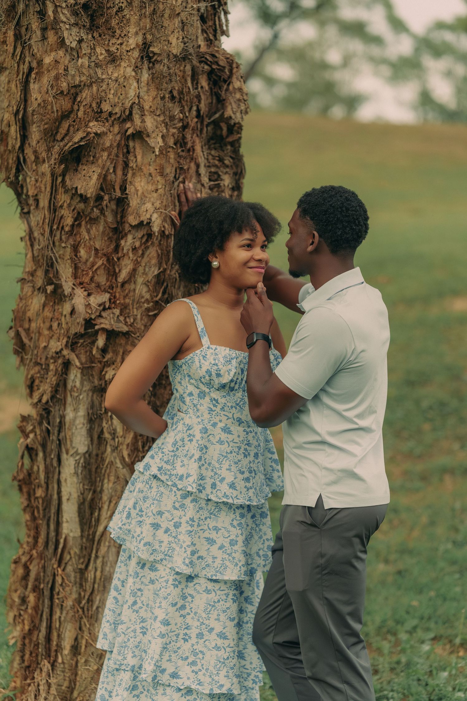 A man tenderly cups a smiling woman's face near a tree trunk in a romantic outdoor park setting.