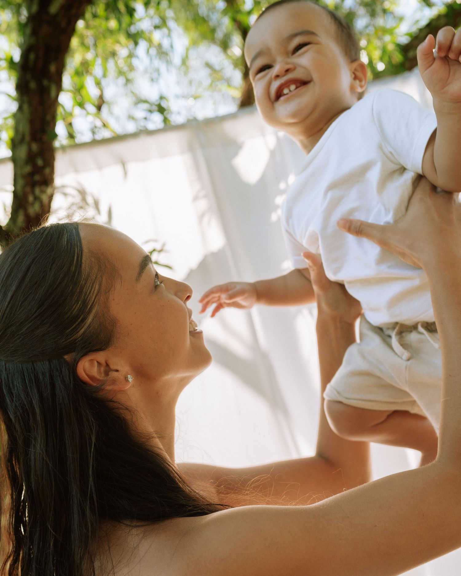 A smiling mother lifts her happy baby boy outdoors in warm sunlight near lush green trees.