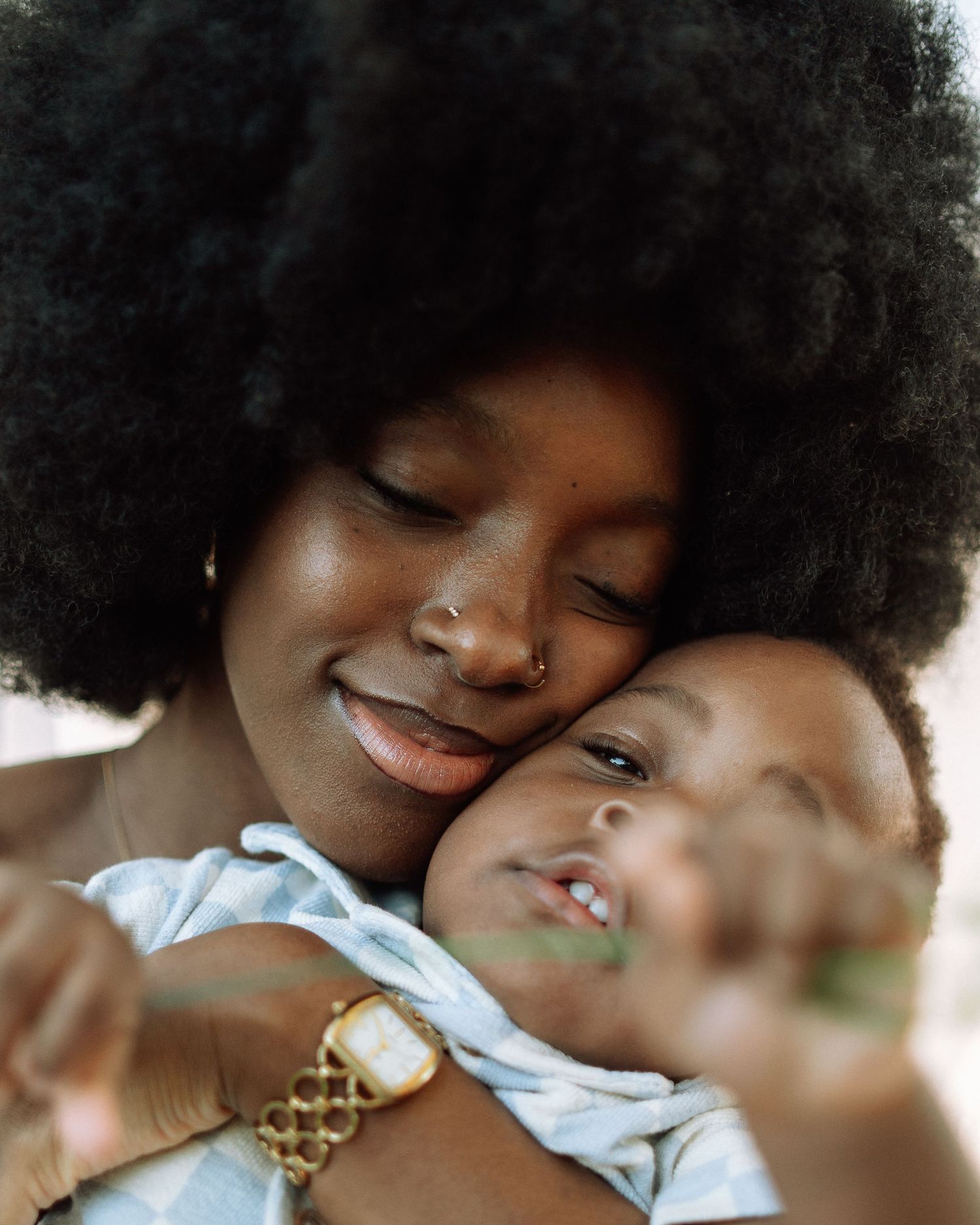 A mother with an afro hairstyle tenderly embraces her baby, both smiling with eyes closed in a warm moment.
