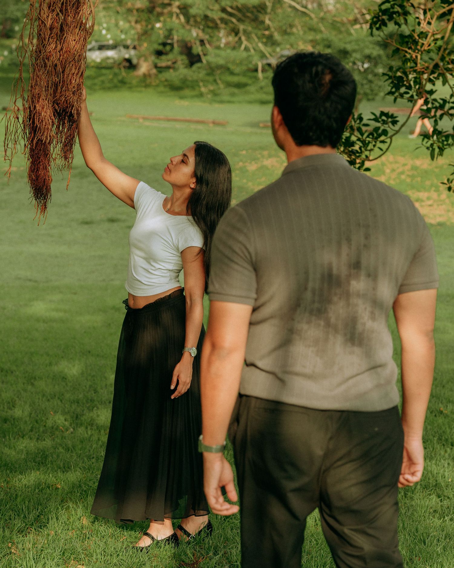 A woman reaches up to touch hanging tree roots while her partner watches in a lush green park.