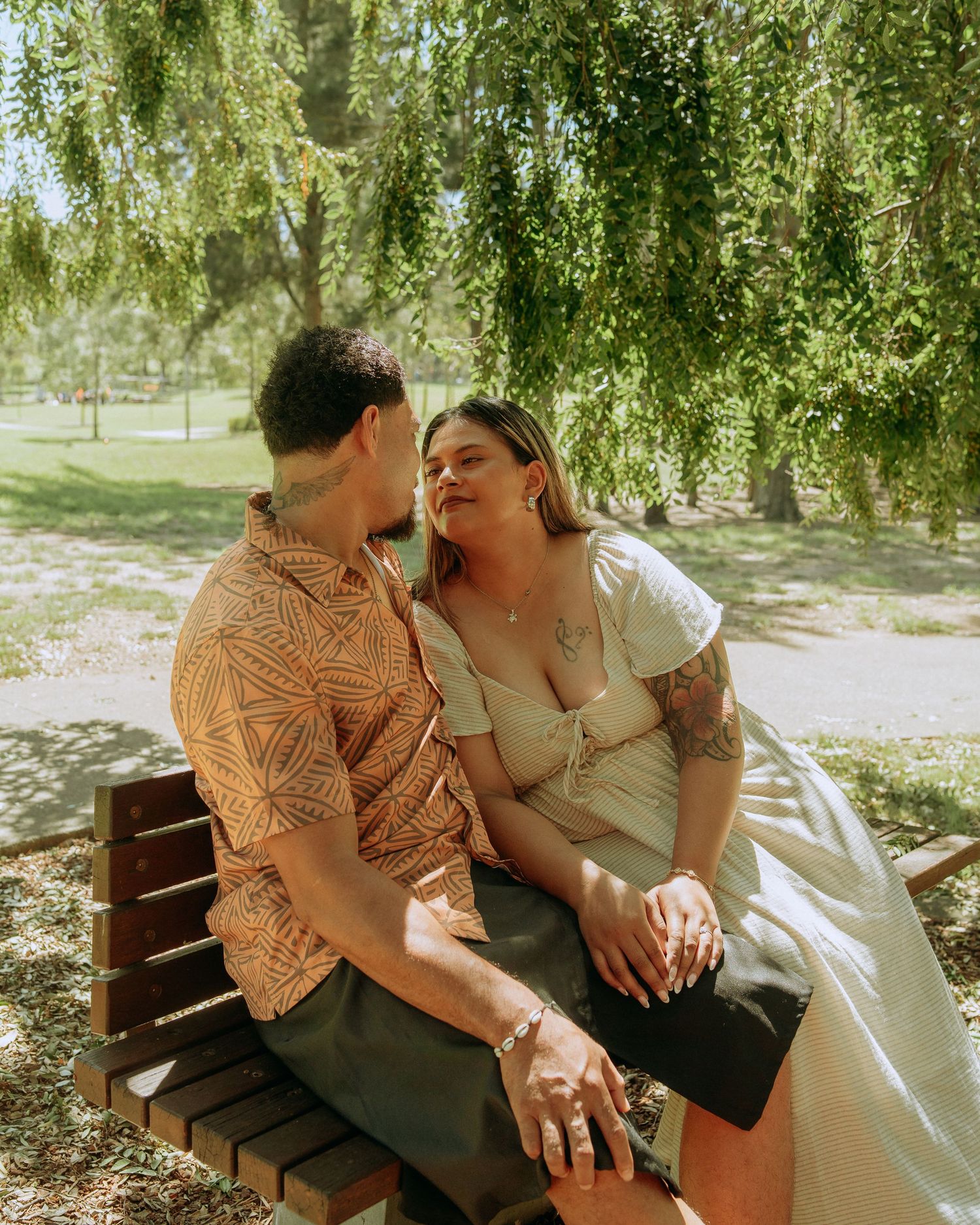 Couple sitting closely together on a wooden bench under weeping willow branches in a sunny park.