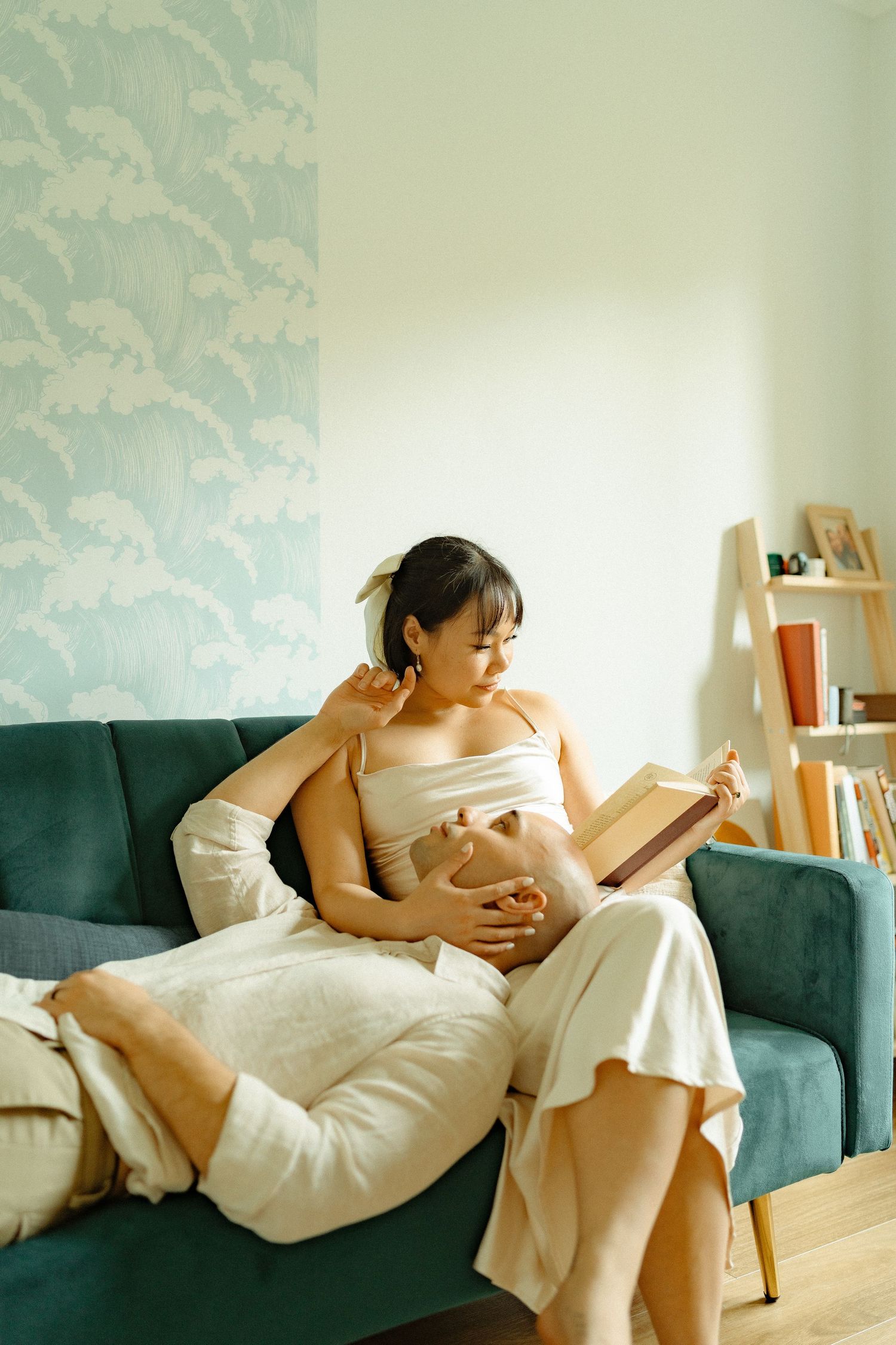 Pregnant woman in white dress reading book on teal sofa, partner embracing her from behind near bookshelf.