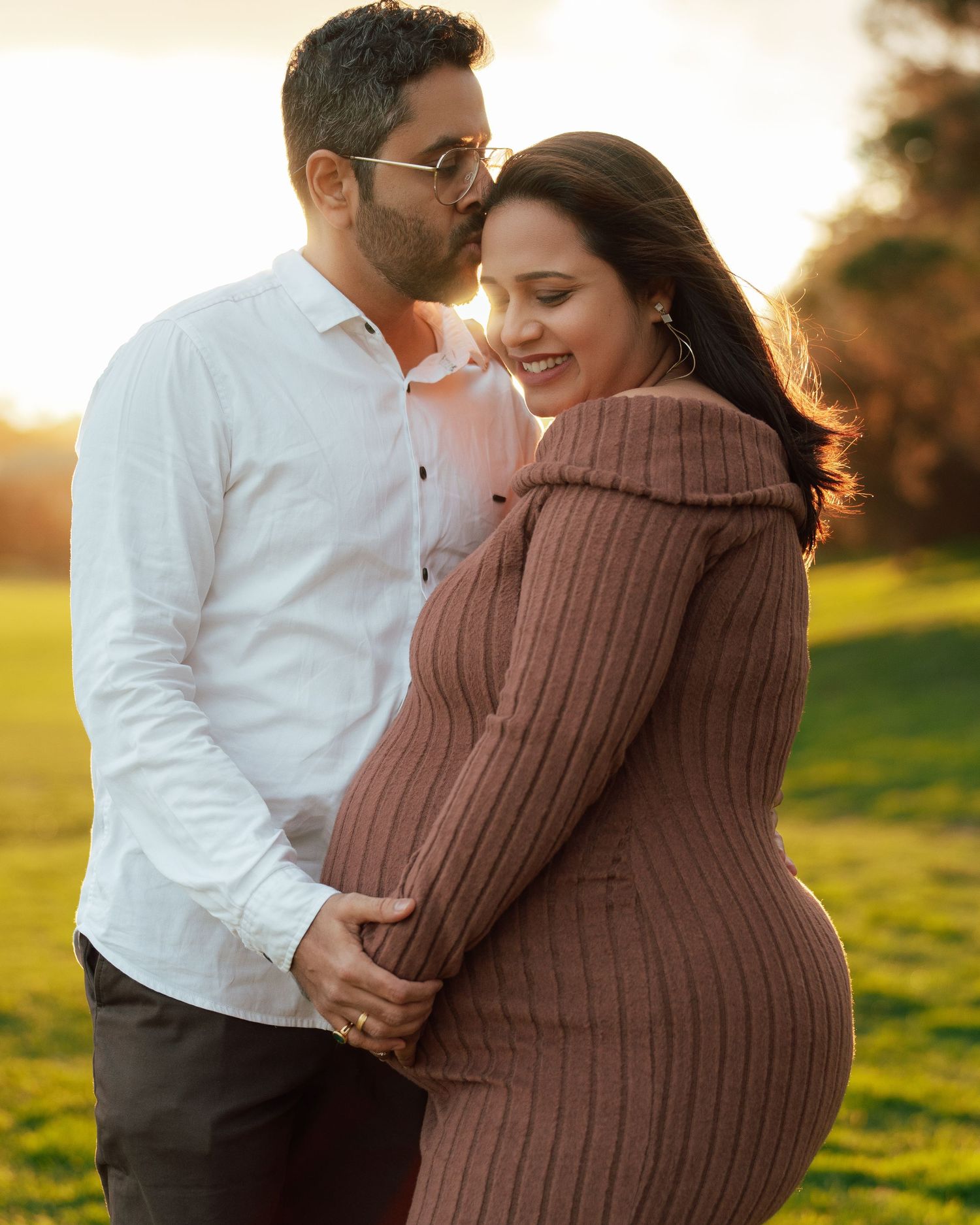 Couple sharing a tender maternity photo outdoors at golden hour, man embracing pregnant woman in brown knit dress.
