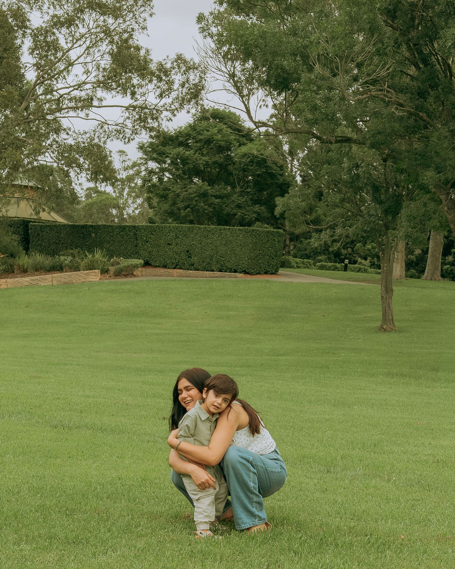 A mother crouches on green grass hugging her young child in a warm embrace in a park setting.