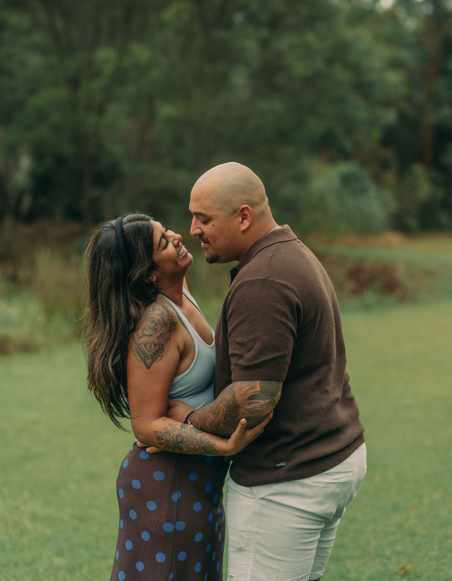 A tattooed couple shares a romantic moment in a lush green park, gazing into each other's eyes.