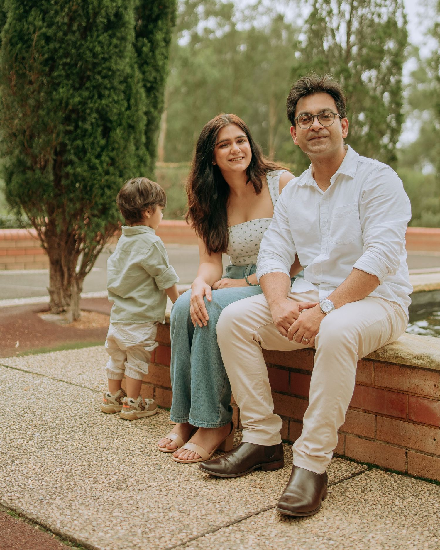 Family of three posing outdoors, parents sitting on brick wall while toddler stands nearby in a lush green park.