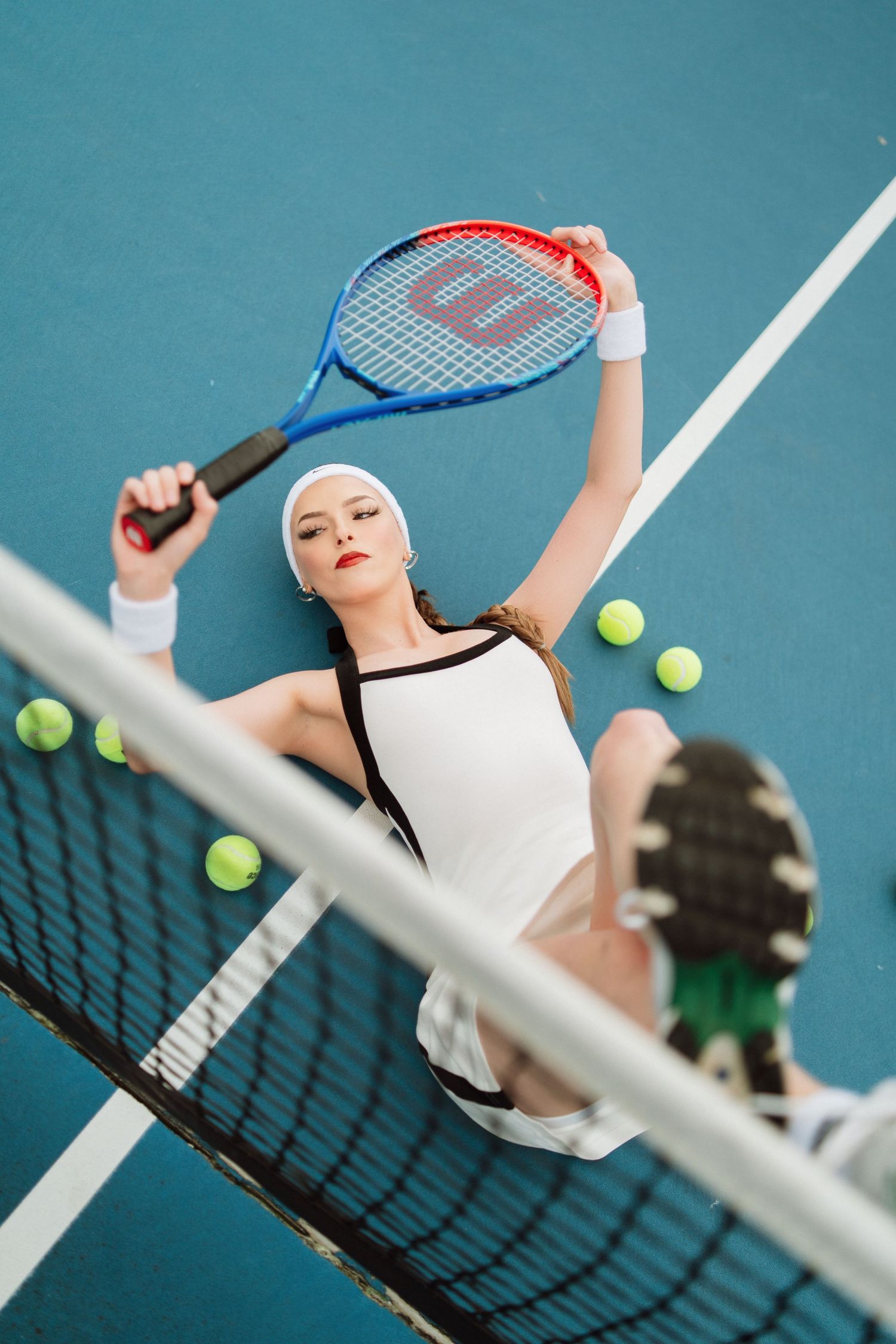 Female tennis player lying on blue court holding Wilson racket with tennis balls nearby.