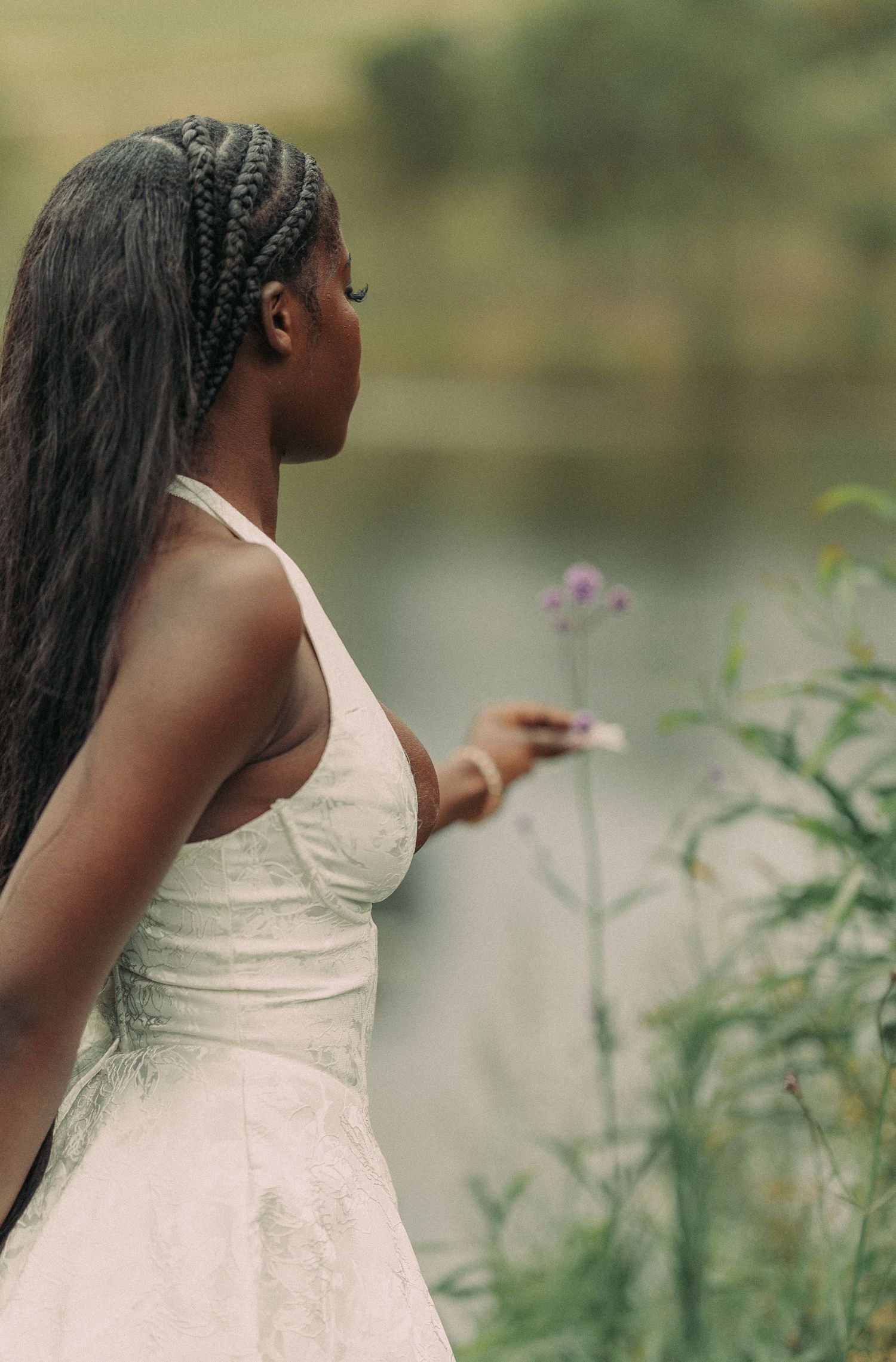 A young Black woman in a white lace halter dress holds a small flower near a serene misty lake.