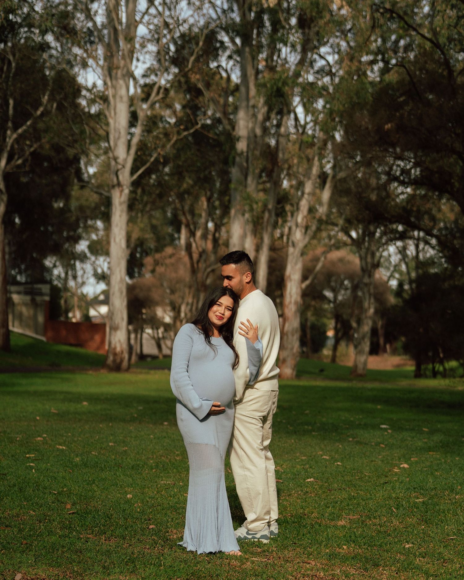 Couple posing for maternity photos in a park, woman in blue dress cradling baby bump, man in cream outfit behind her.