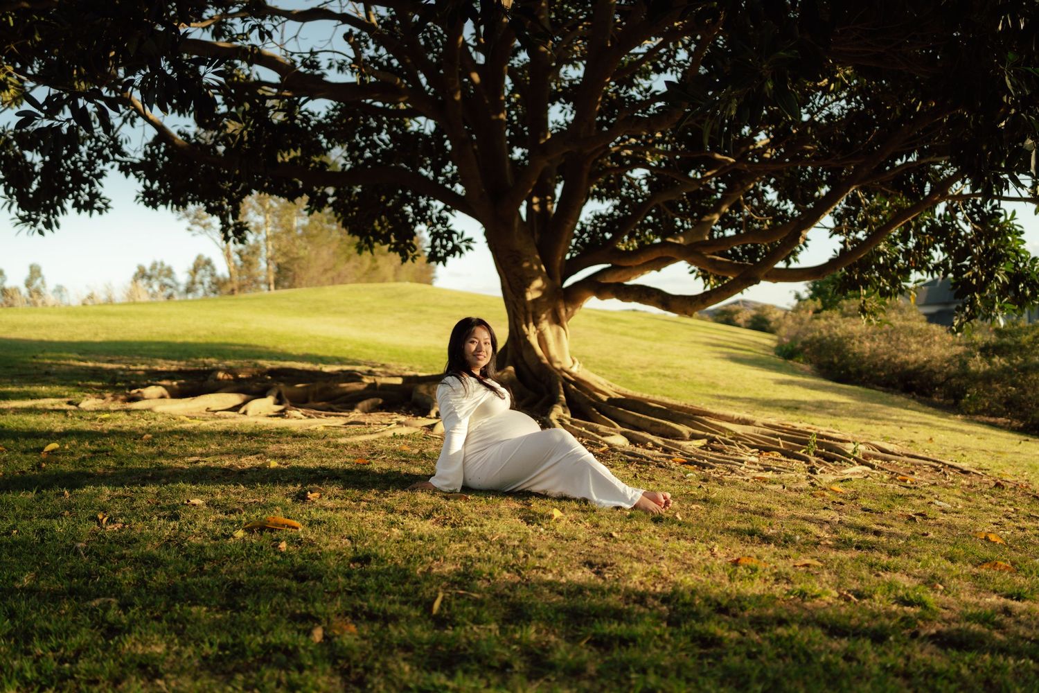 Woman in white dress sitting under a large tree on green grass in golden hour sunlight outdoors.