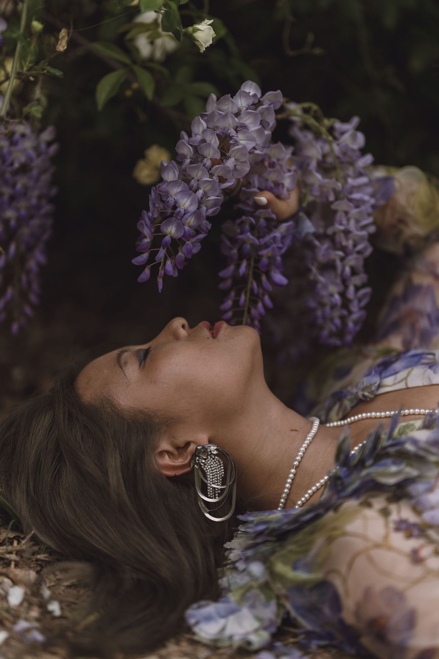 Woman lying among purple wisteria flowers, wearing floral dress with pearl necklace and earrings.