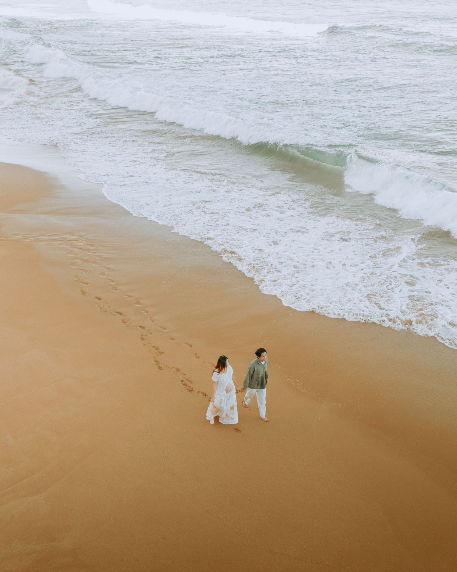 Aerial view of couple walking hand-in-hand along a sandy beach with rolling ocean waves.