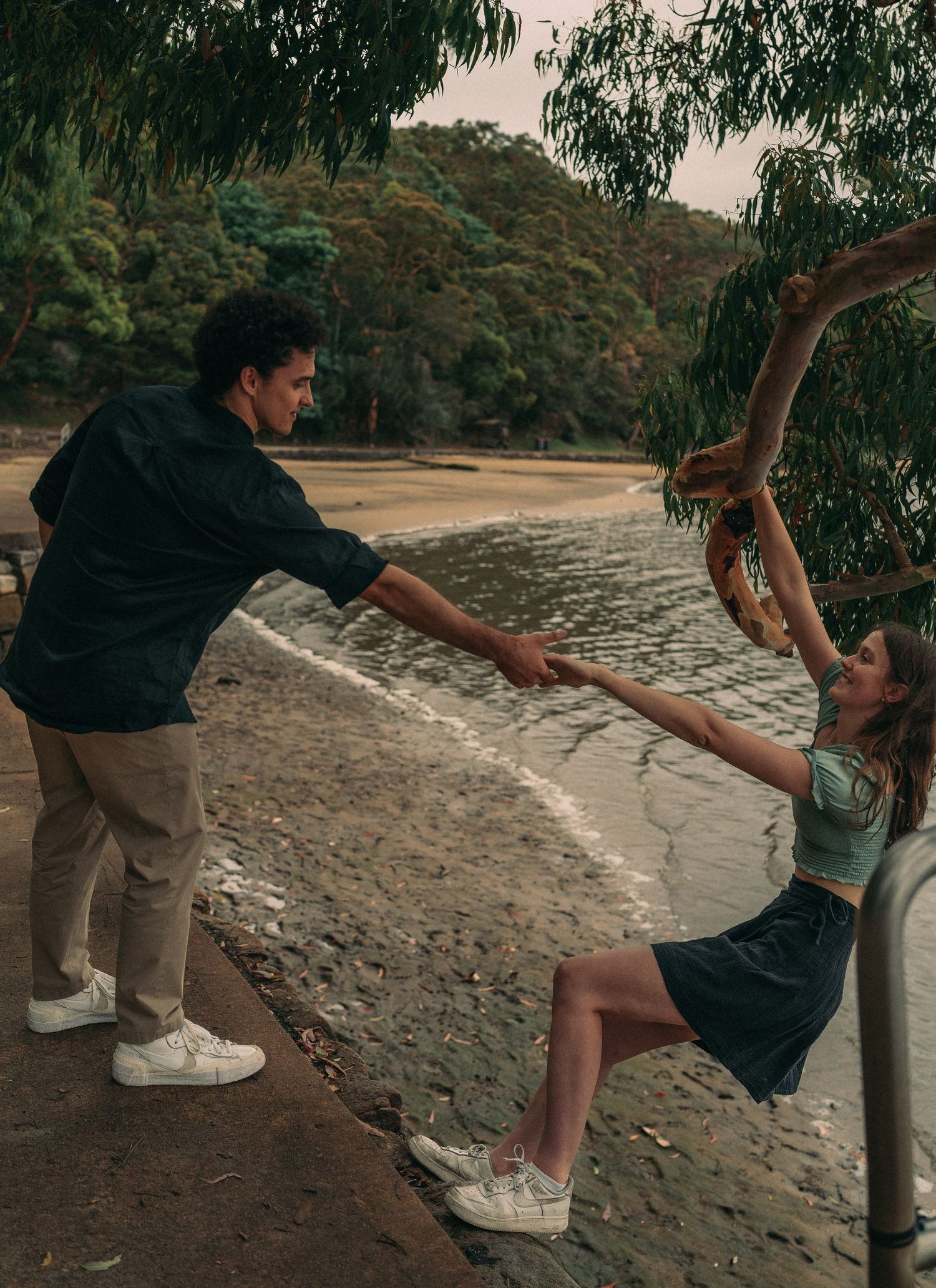 A couple playfully holding hands near a scenic beach with a tree branch overhead.