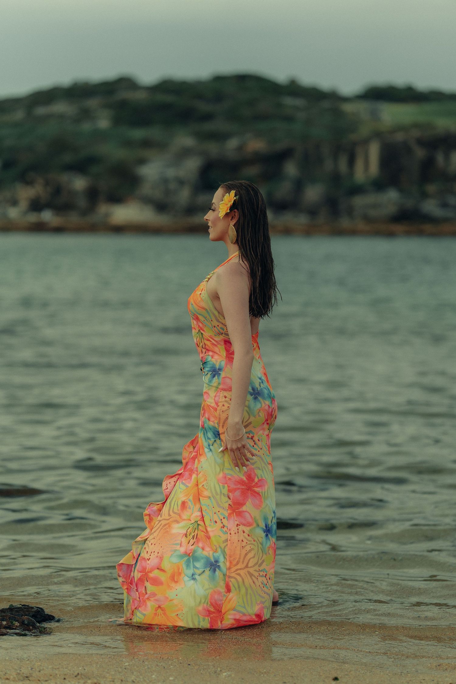 Woman in colorful floral maxi dress standing in shallow ocean water at dusk with rocky hills in background.