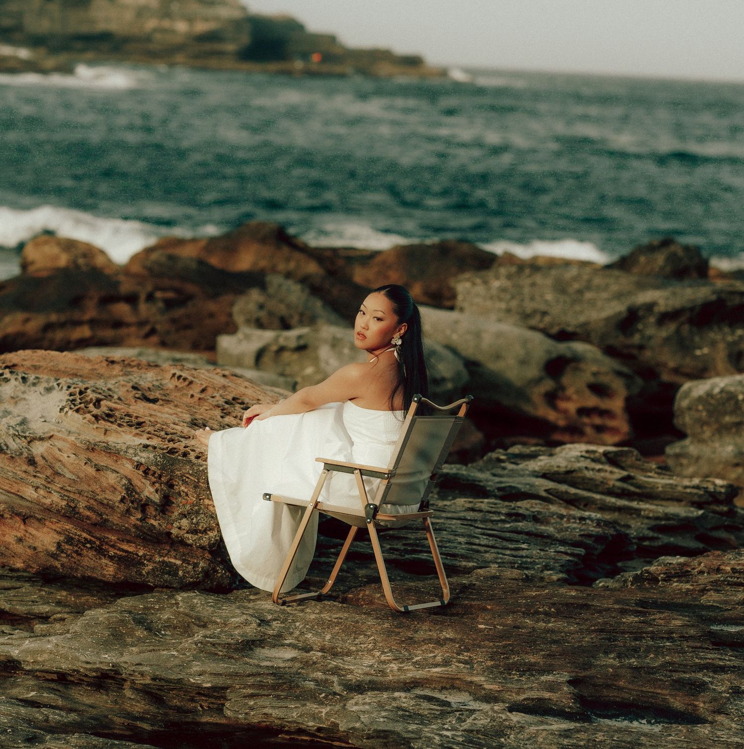 Woman in elegant white dress seated on folding chair atop coastal rocks with turquoise ocean backdrop.