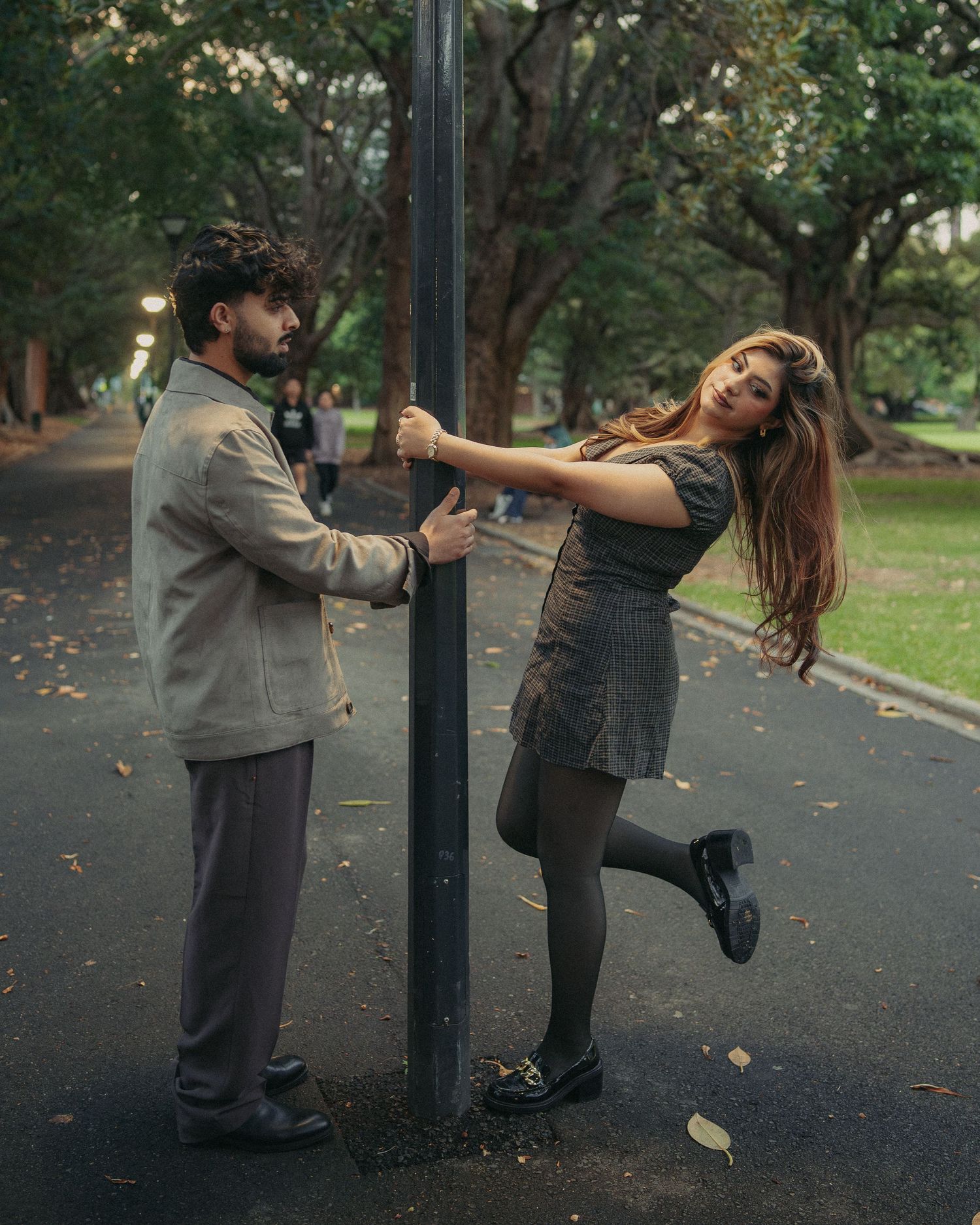Playful couple posing on opposite sides of a lamp post in a lush green park at dusk.
