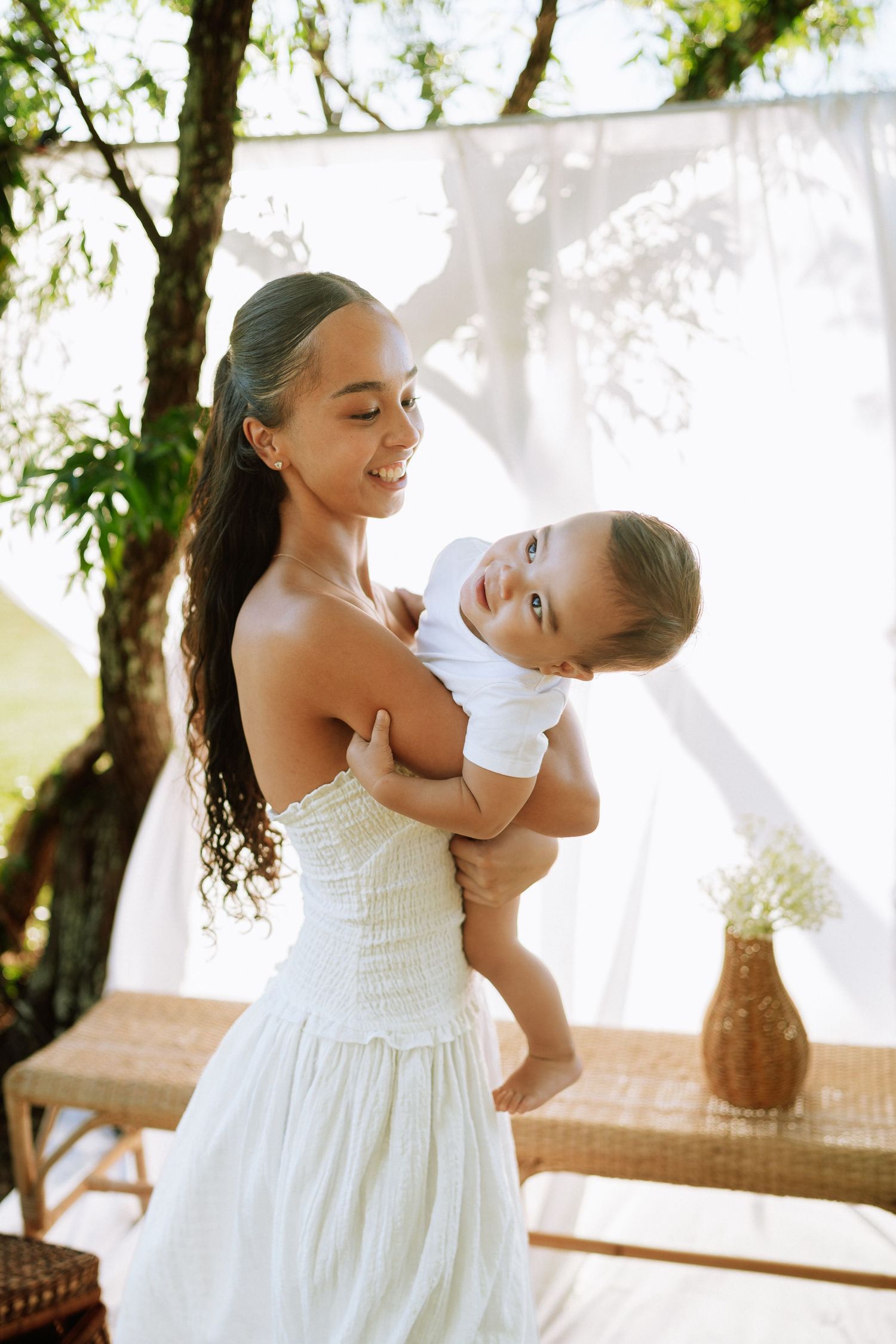A smiling woman in a white dress holds a happy baby outdoors near sheer white curtains.