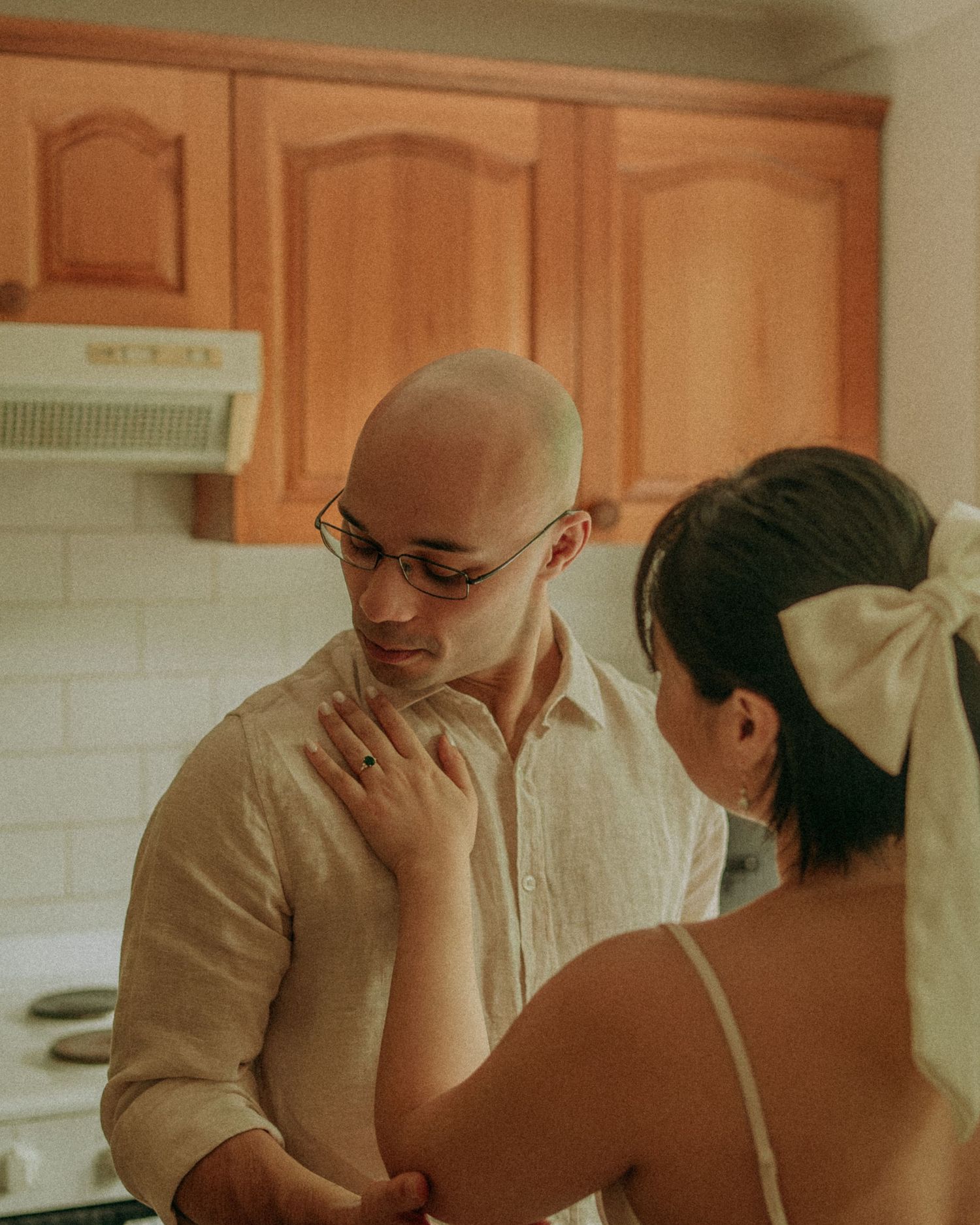 Woman in slip dress gently touches bald mans chest in cozy kitchen, sharing an intimate tender moment.
