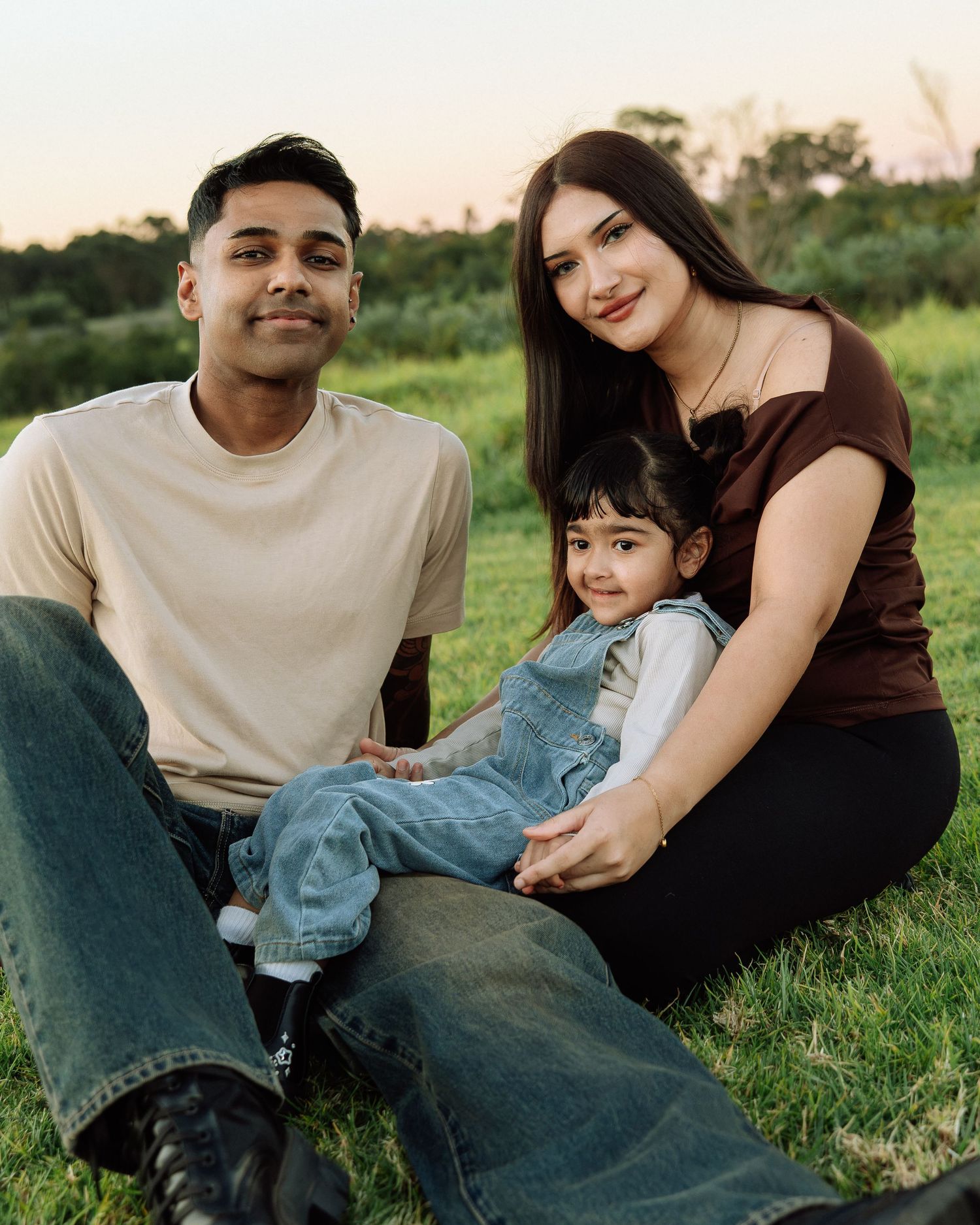 Young family of three sitting on grass at sunset, parents smiling with toddler in denim overalls.