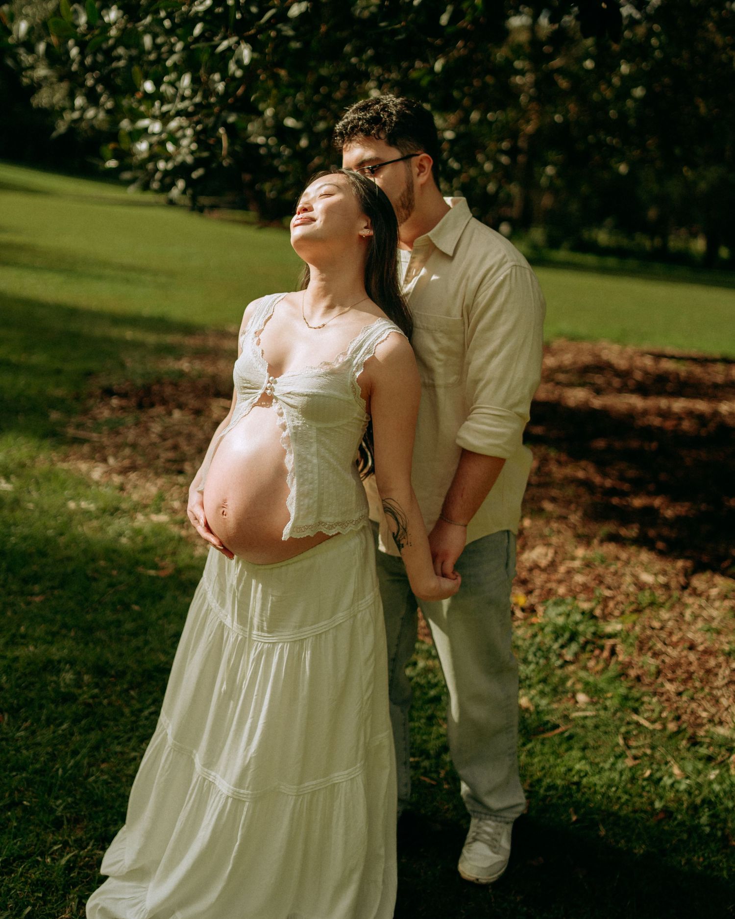 Couple in sunlit park, pregnant woman in white dress cradling baby bump, partner embracing her from behind.