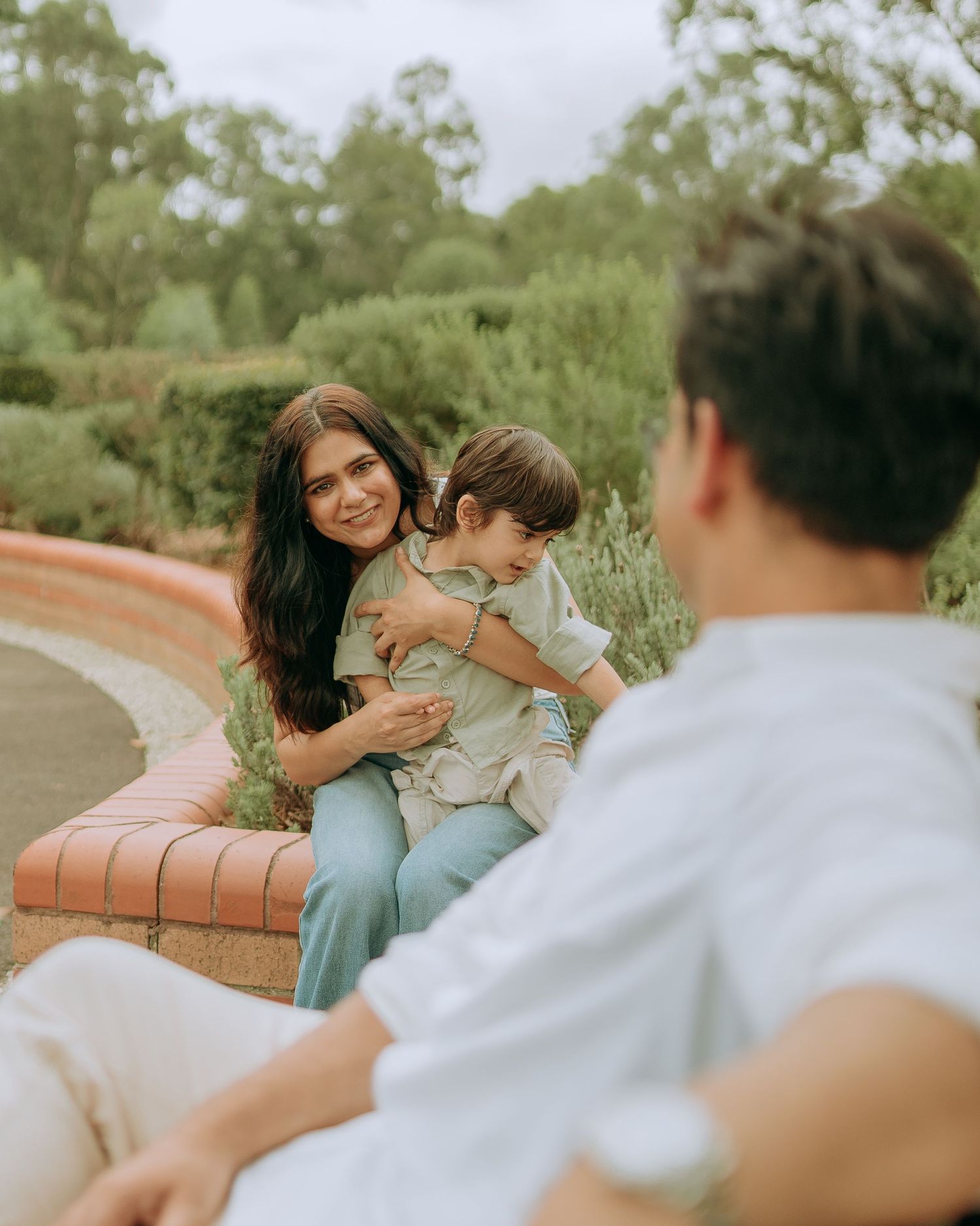 Mother sitting on curved brick bench holding toddler son, smiling at father in a lush garden setting.