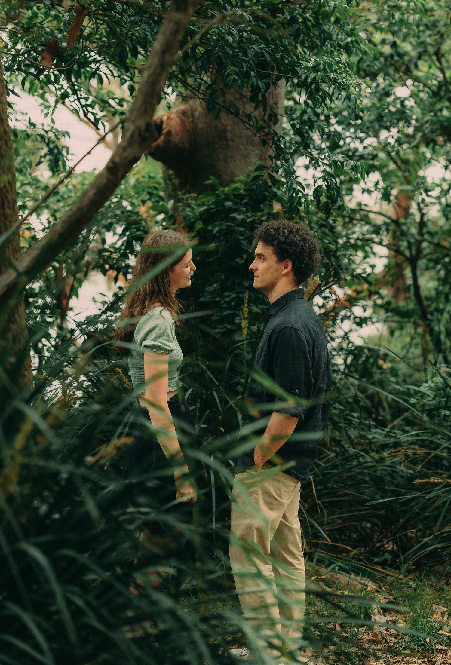Young couple facing each other in a lush green forest, sharing an intimate moment among the trees.