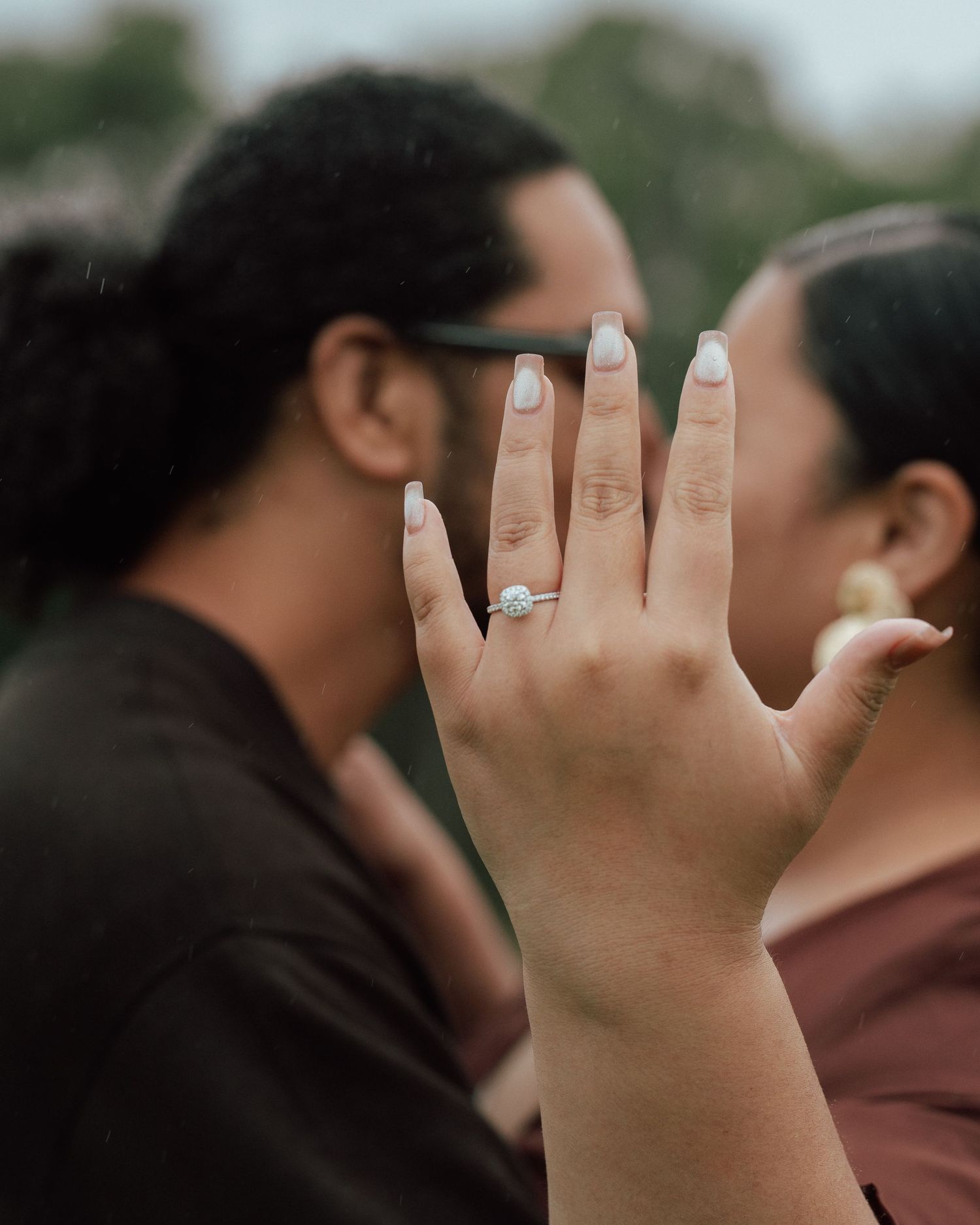 A woman proudly displays her diamond engagement ring while kissing her partner outdoors.
