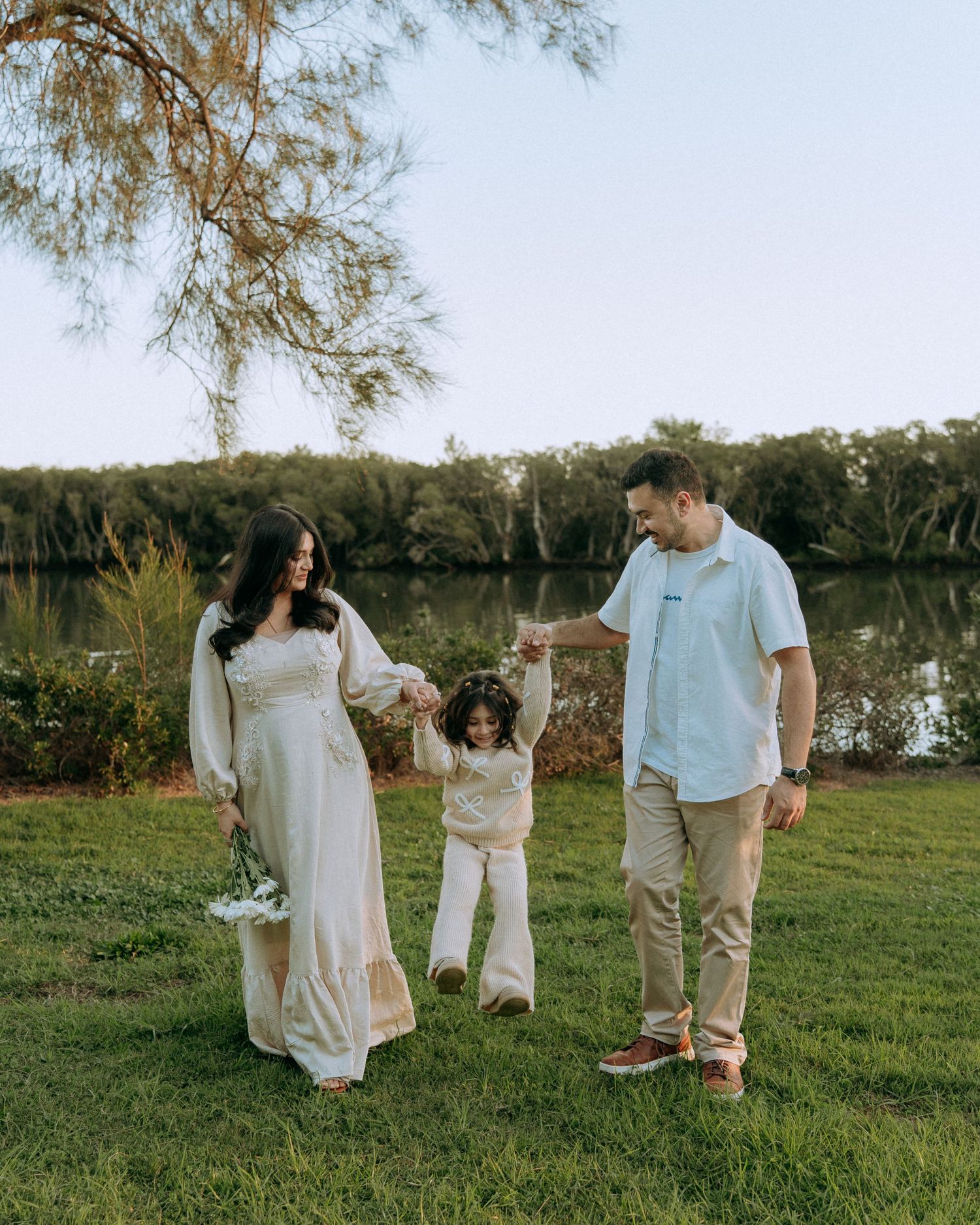 Happy parents swing their young child between them while walking on green grass near a peaceful lakeside setting.