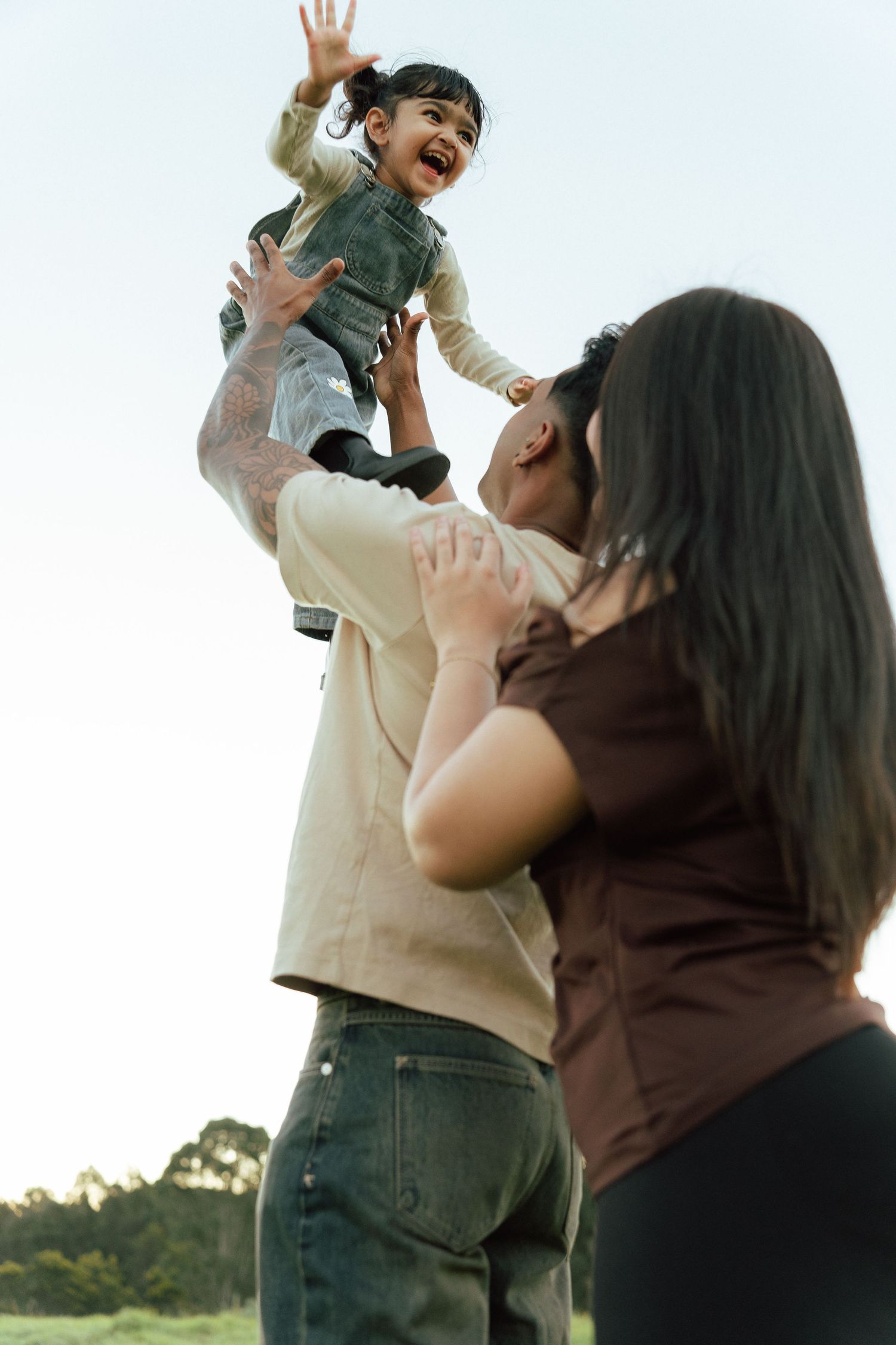 Parents joyfully lifting their laughing child into the air outdoors, sharing a happy family moment in a park.