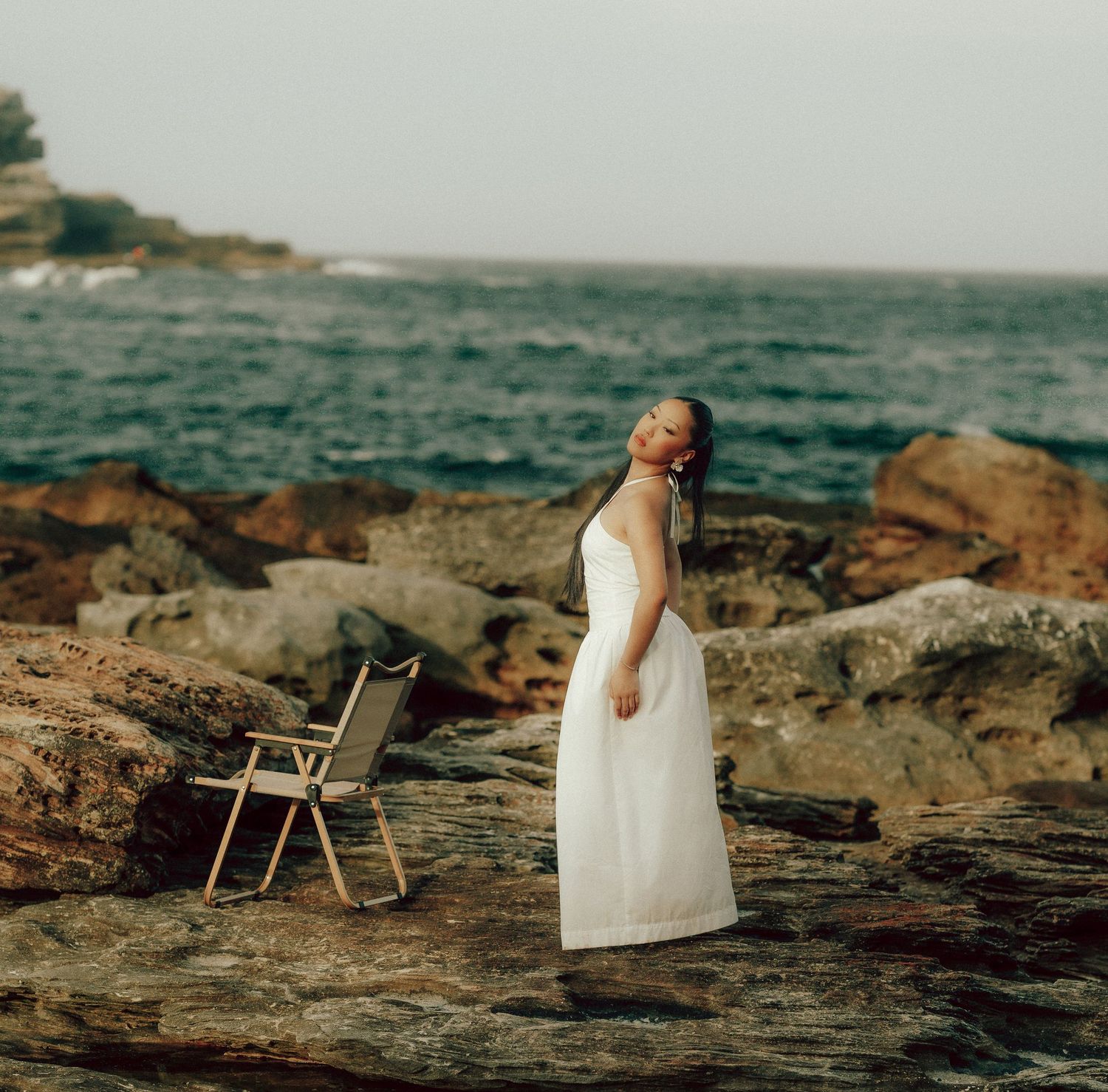 Woman in flowing white dress standing on rocky shore with calm sea and misty horizon in background.