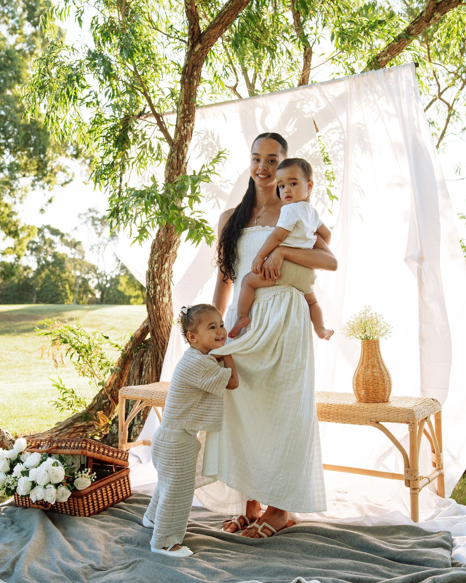 Woman in white dress holds baby outdoors under tree with draped fabric backdrop and rattan bench, while toddler hugs her leg.